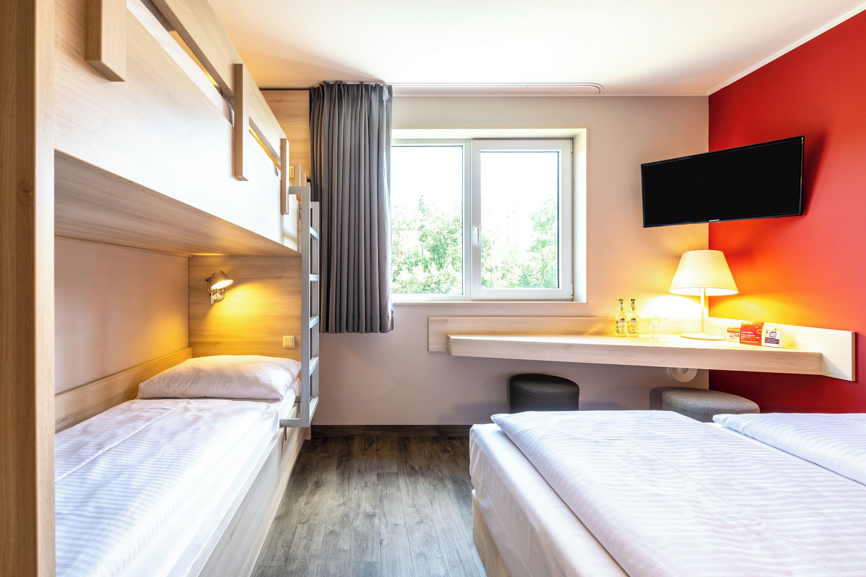 Habitación de hotel con dos camas, una ventana con cortinas, un televisor montado, una mesita de noche con lámpara y un taburete pequeño.