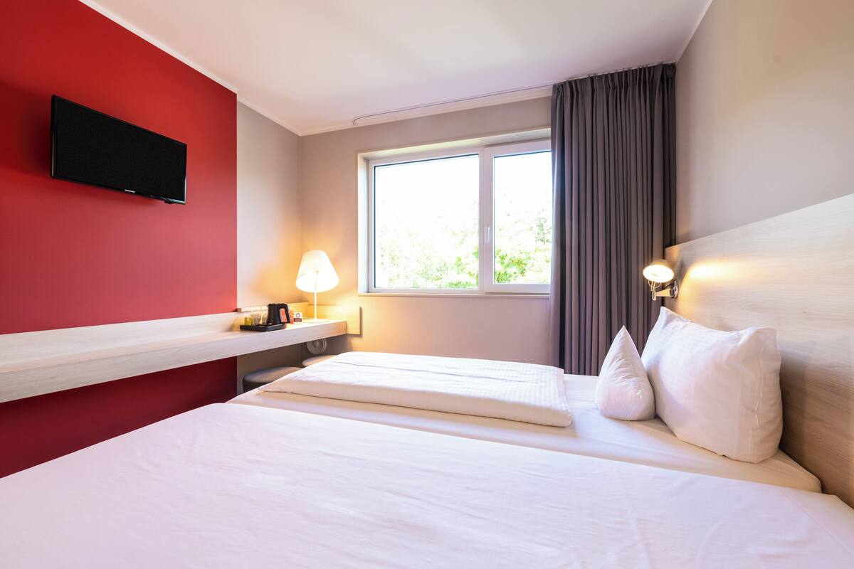 A hotel room with a bed, a TV mounted on the wall, a window with curtains, and a bedside lamp.