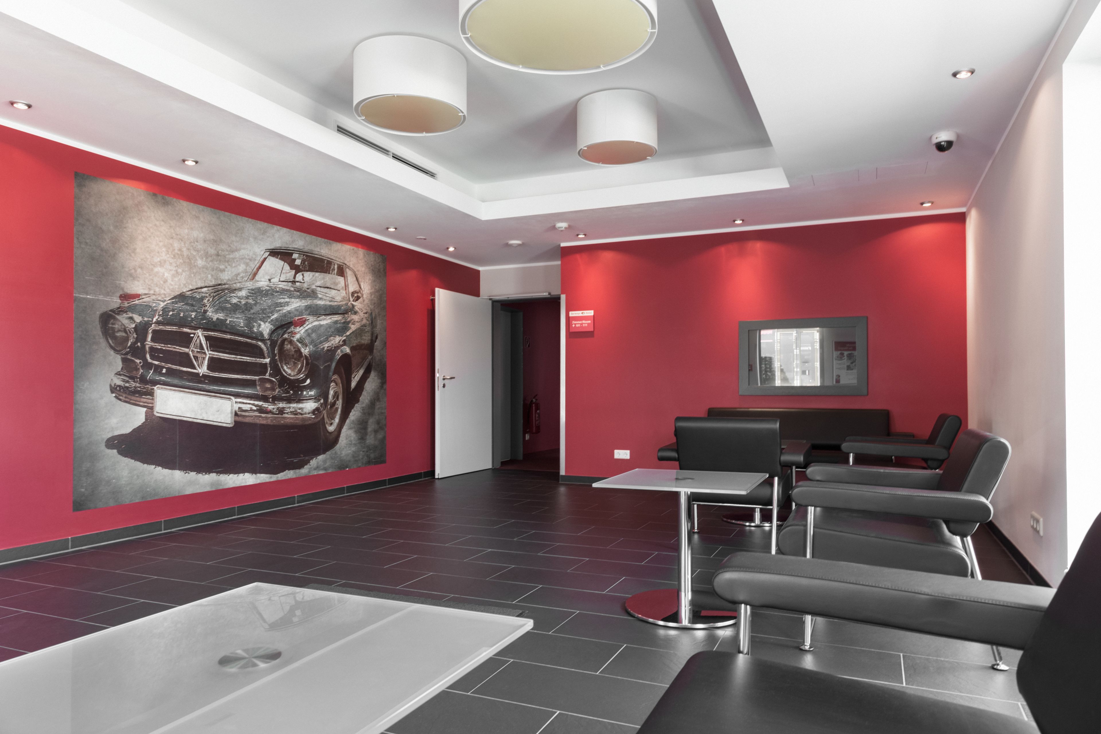 Un área de espera moderna con un gran mural de un coche clásico, paredes rojas y mobiliario contemporáneo.