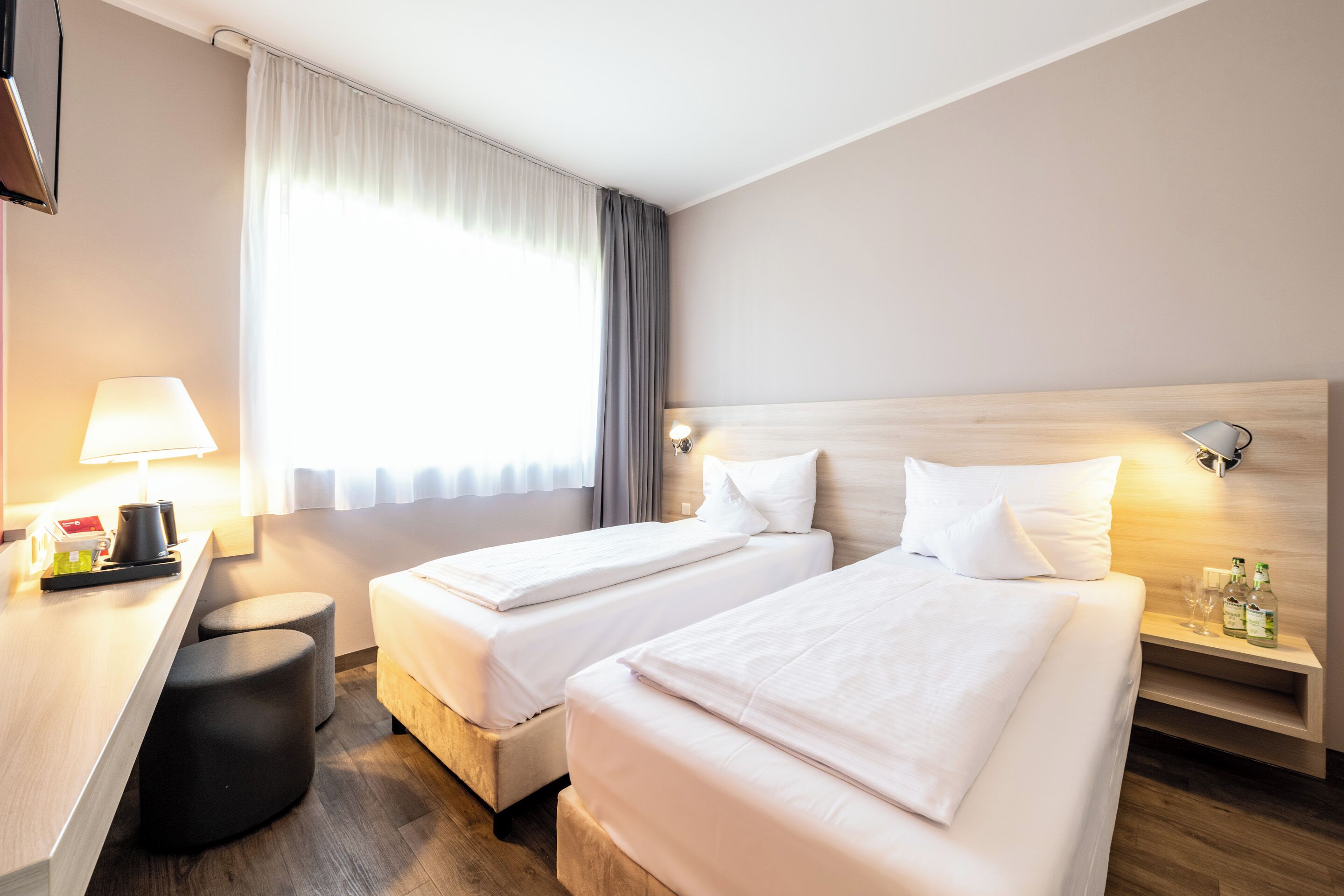 Habitación de hotel con dos camas, un escritorio, una lámpara y una mesita de noche con botellas.