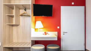 A room with a wooden wardrobe, a mounted television, a white bedside table with a lamp, and two grey stools. - Serways Hotel Nürnberg Feucht Ost (Schwarzenbruck)