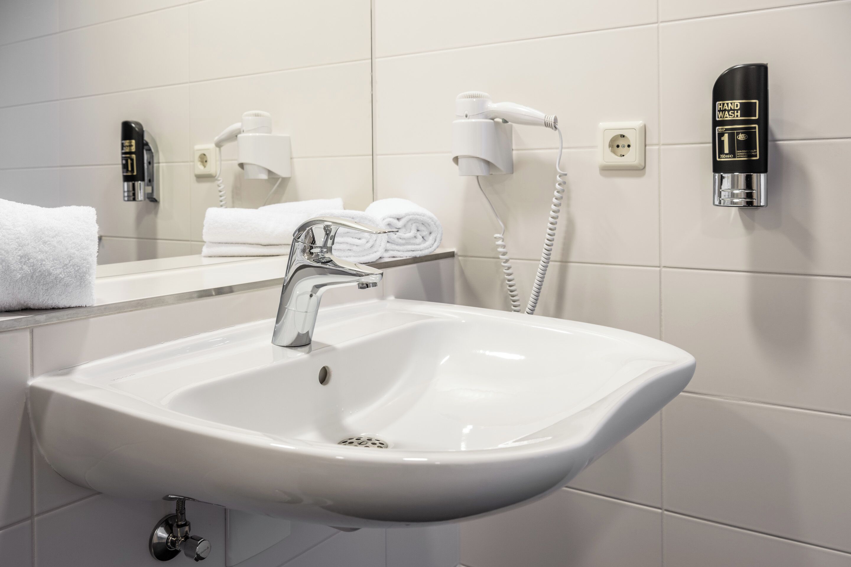 A modern bathroom with a white sink, a mirror, a hairdryer, and a hand wash station.