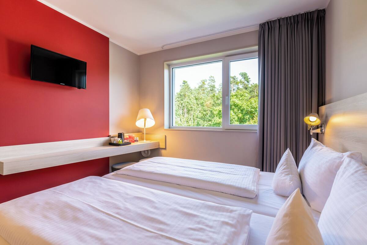 A hotel room with two beds, a TV mounted on the wall, a window with curtains, and a bedside table with a lamp.