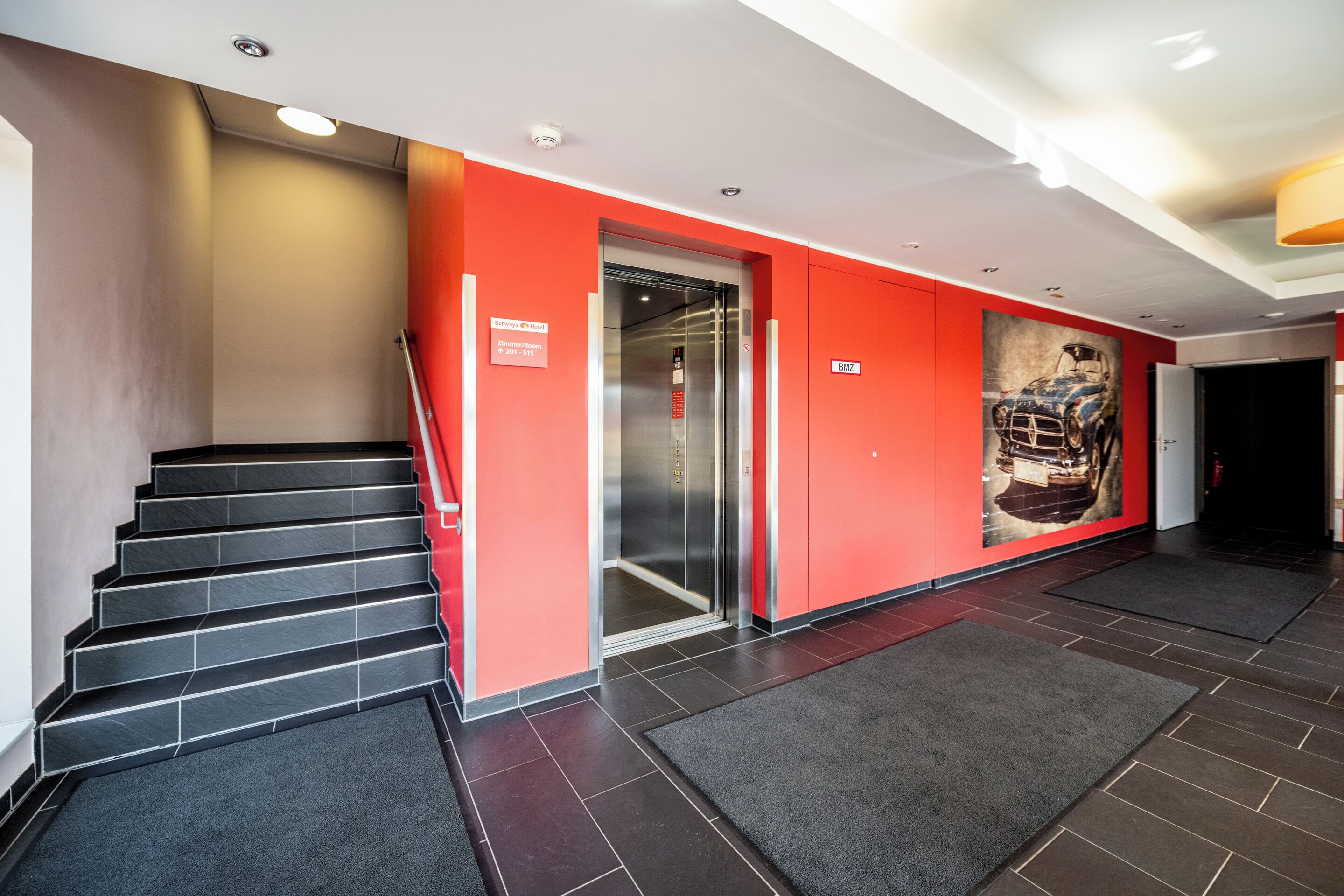 A modern hotel lobby with a red accent wall, a staircase, and a large artwork of a car.