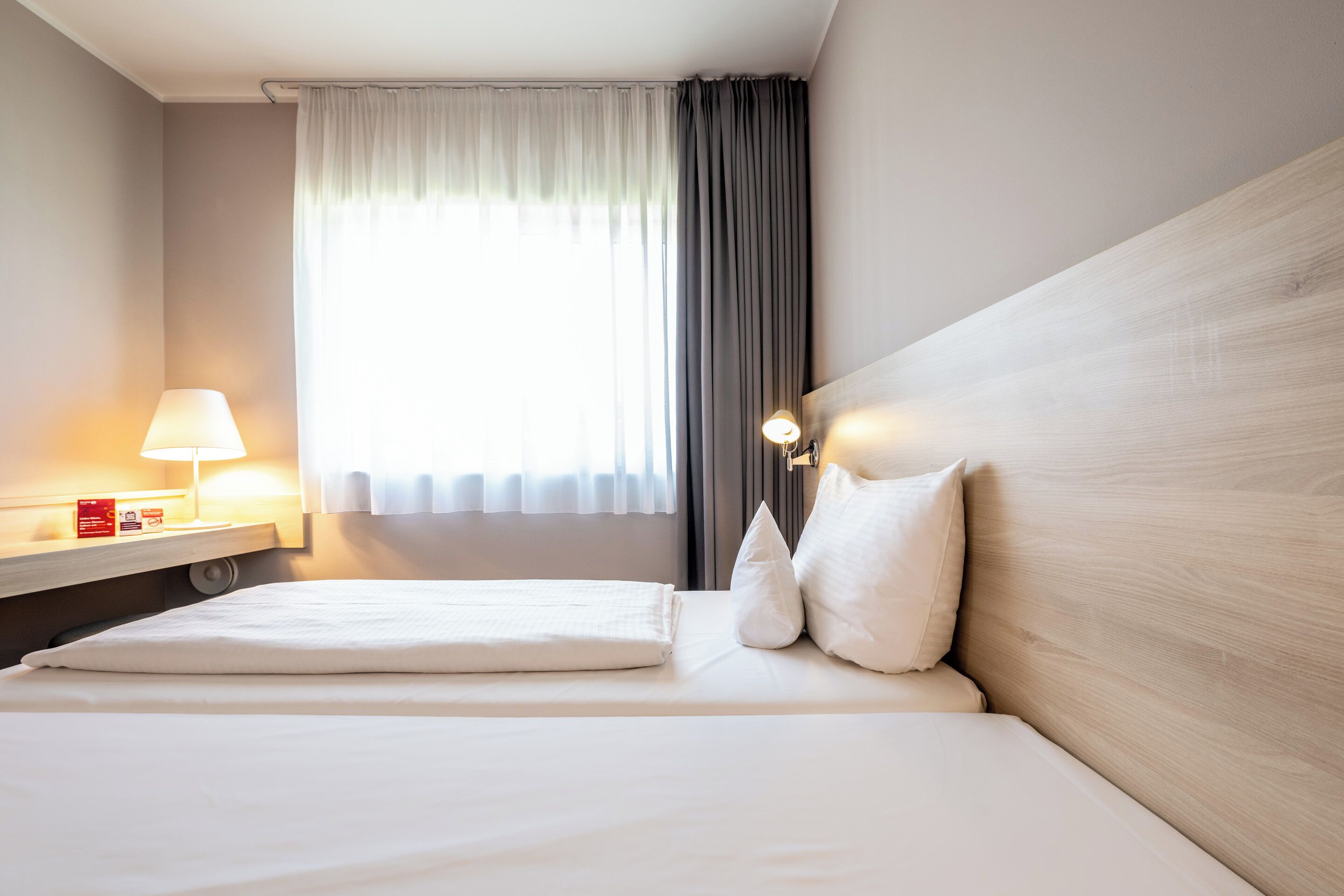 Una cama bien hecha con sábanas blancas, una lámpara de noche y un ventanal con cortinas.