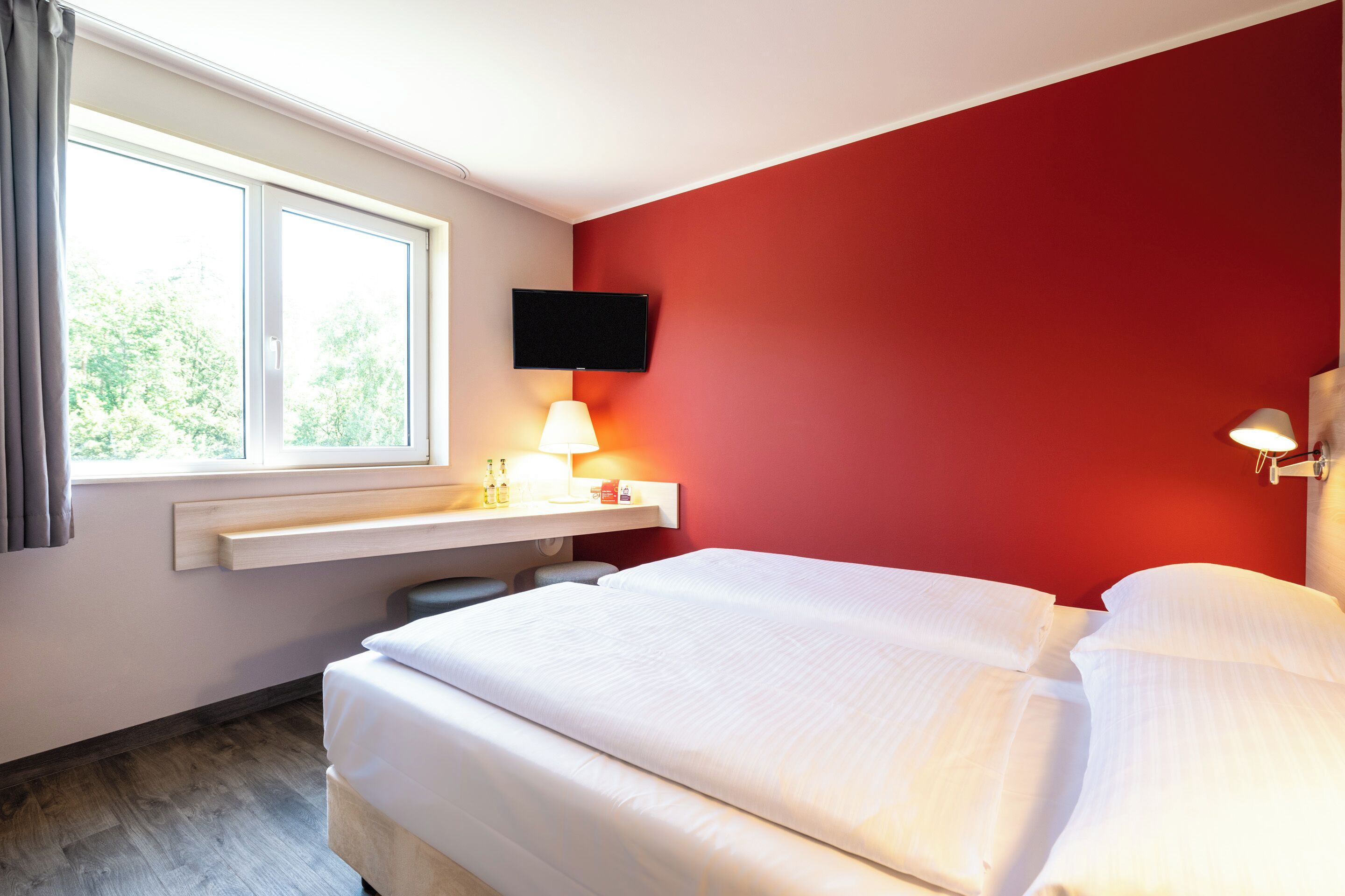 Une chambre d’hôtel avec un lit, un téléviseur fixé au mur, une table de chevet avec une lampe et une fenêtre avec des rideaux.