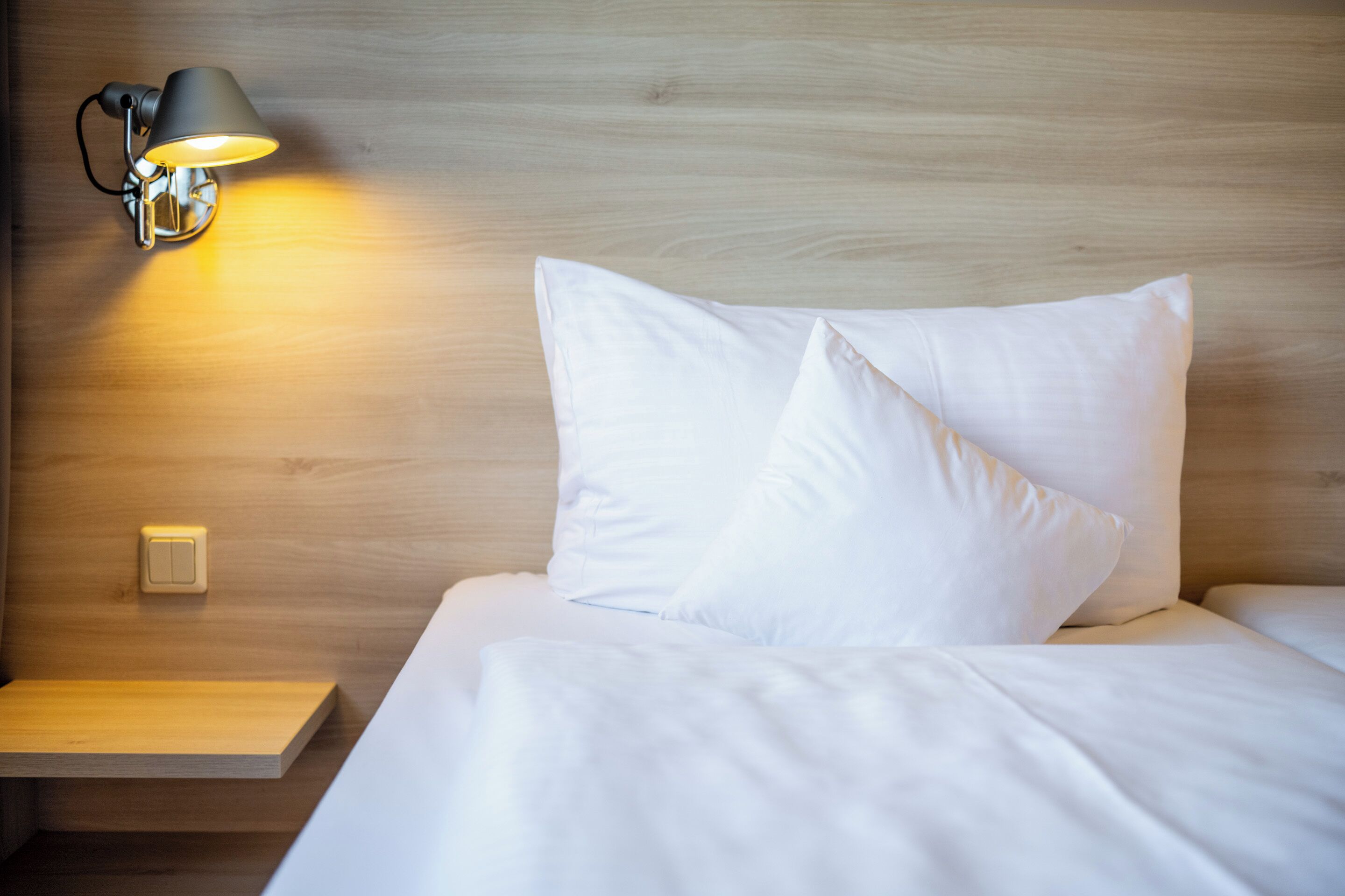 A neatly made bed with white linens and pillows, a bedside table with a lamp, and a wooden wall.