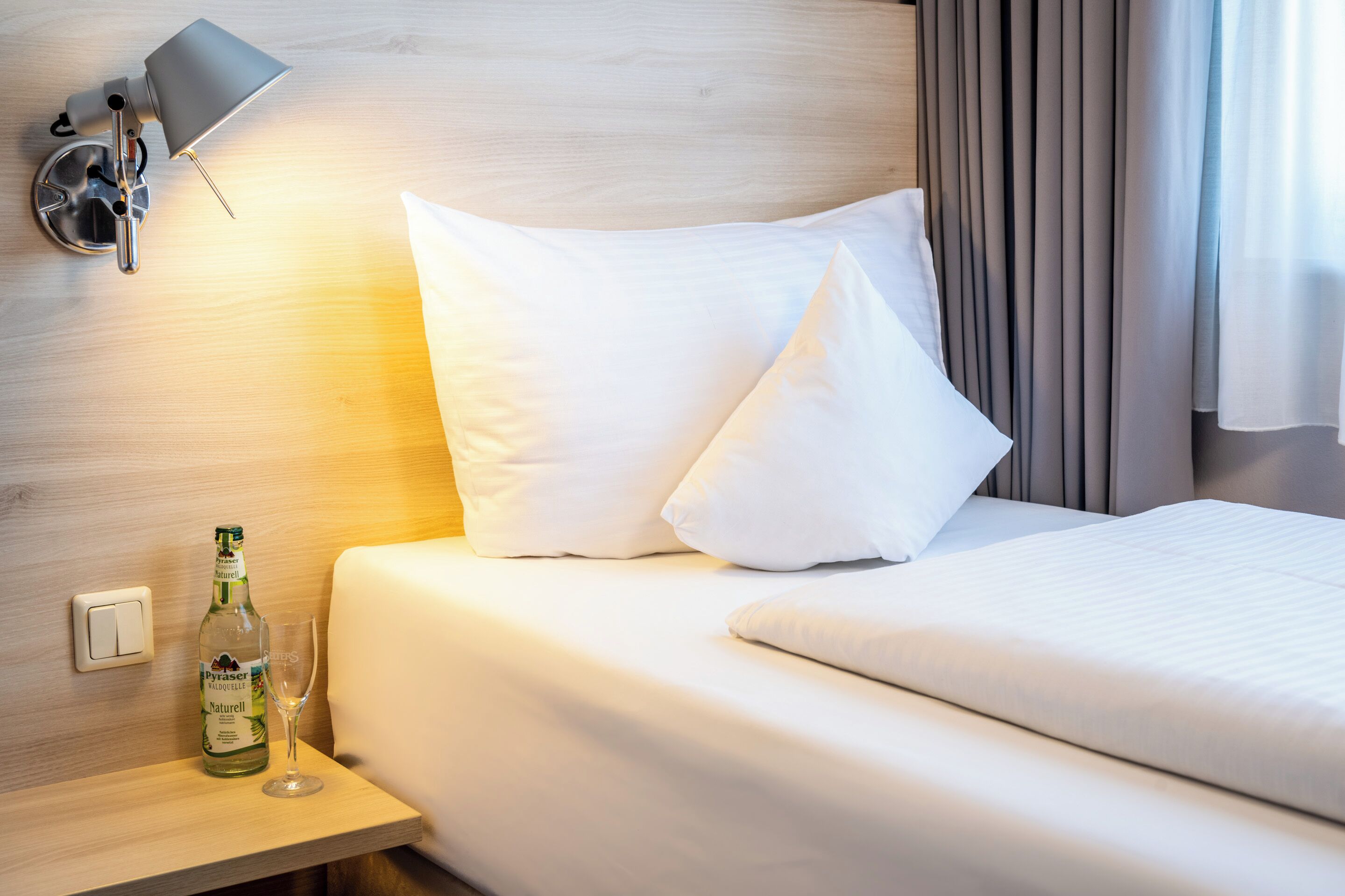 A hotel room with a neatly made bed, a bottle of Pyrazer Naturell on a bedside table, and a lamp on the wall.