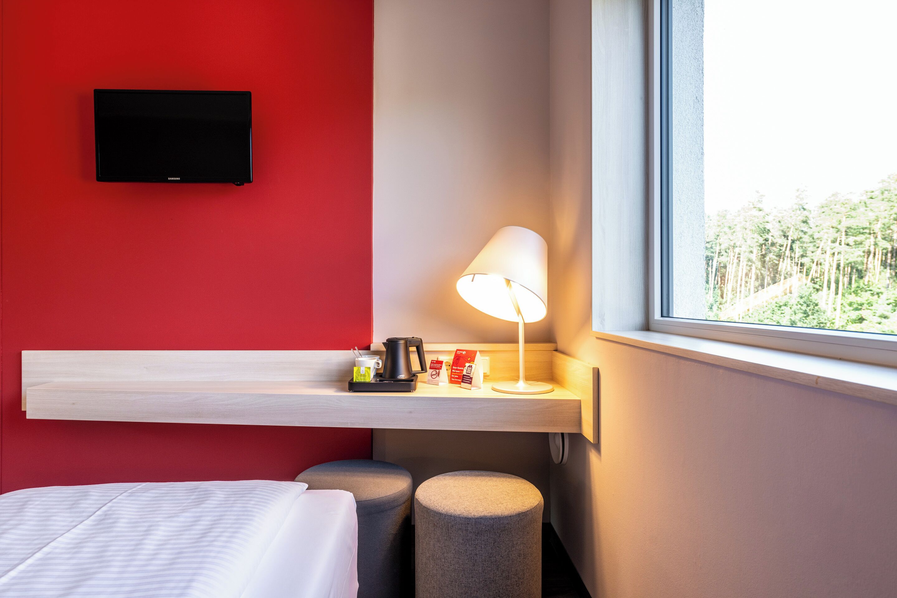 Une chambre d’hôtel avec un lit, un téléviseur fixé au mur, une table de chevet avec une lampe et une fenêtre donnant sur des arbres.