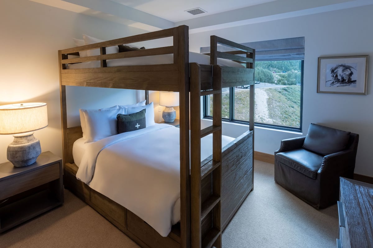 A bunk bed with a wooden frame, a bedside table with a lamp, a chair, and a window with a view of the outdoors.