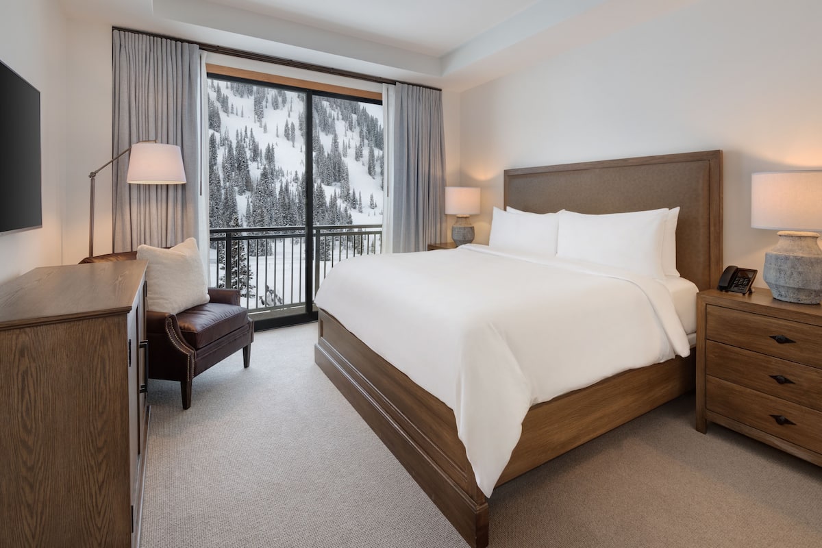 A hotel room with a large bed, a desk, a chair, a nightstand, and a view of a snowy mountain.