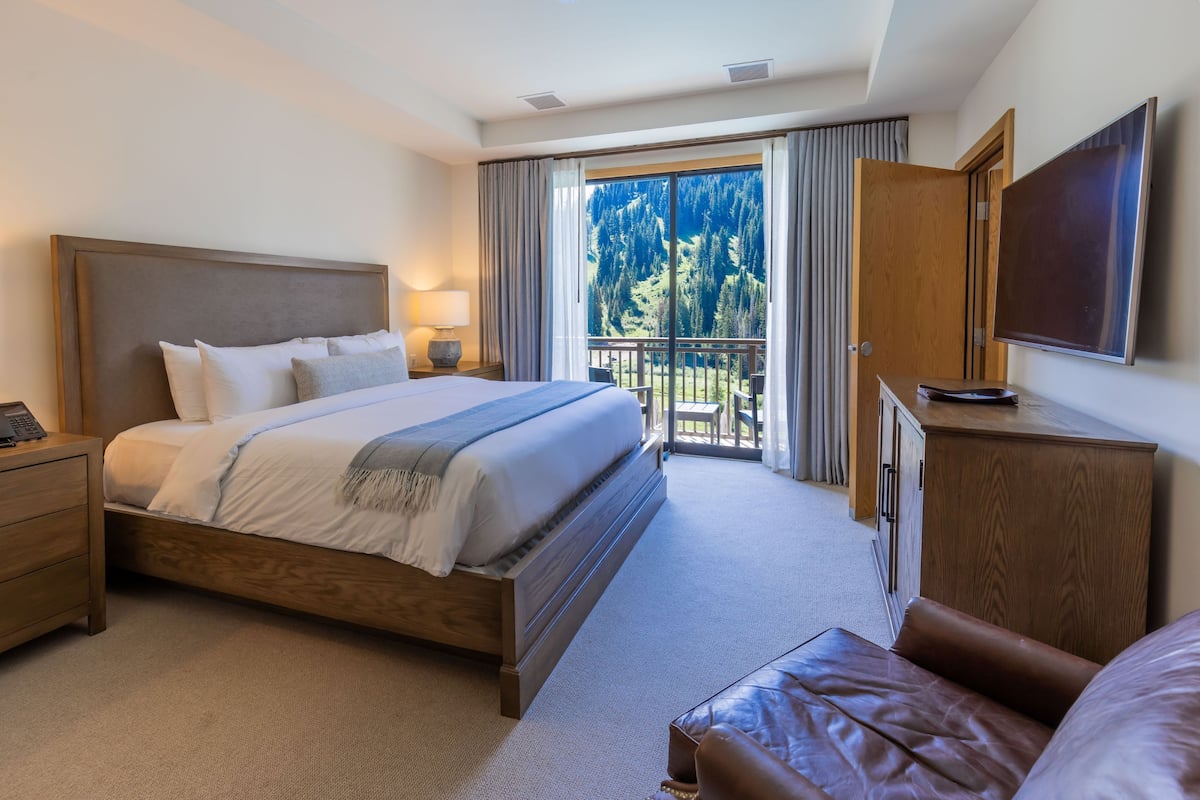 A hotel room with a large bed, a wooden nightstand, a TV, and a balcony with a view of trees.