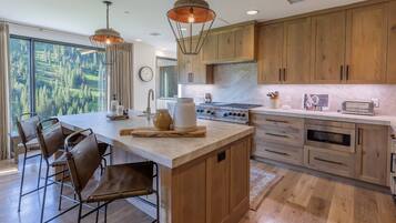 A modern kitchen with wooden cabinets, a large island with bar stools, a stove, and a view of a forest through large windows.