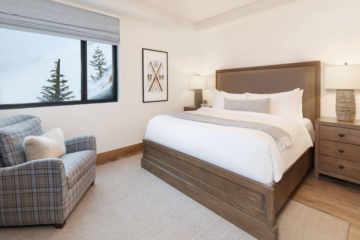 A bedroom with a large bed, a chair, a nightstand, a lamp, and a window with a view of trees.