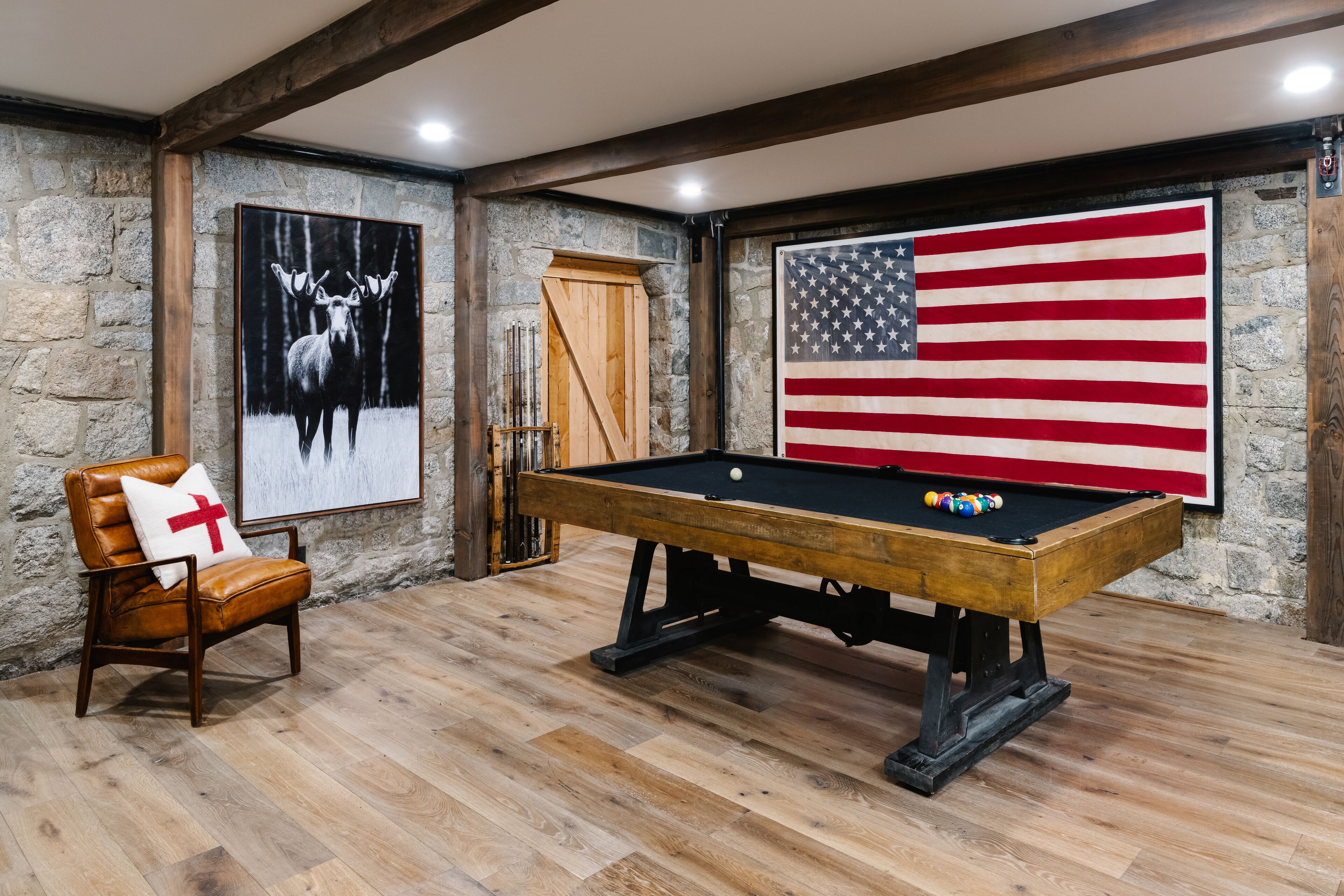 Une table de billard dans une pièce aux murs de pierre, une chaise en bois avec un coussin et un grand drapeau américain.
