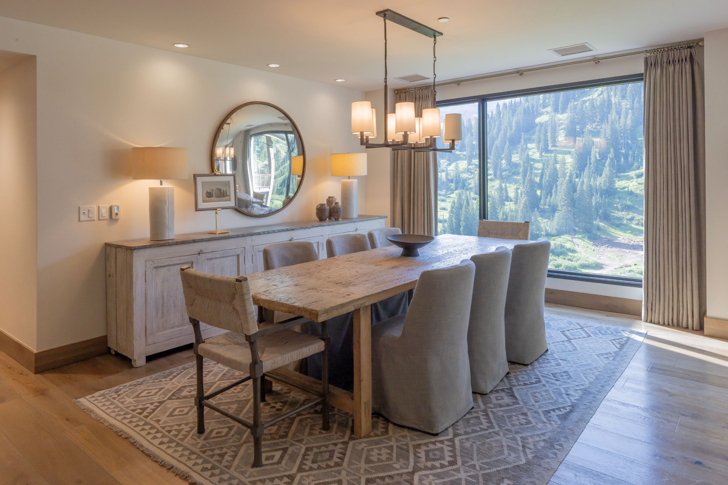A modern dining area with a large table, chairs, a chandelier, a mirror, and a view of a mountainous landscape through a large window.