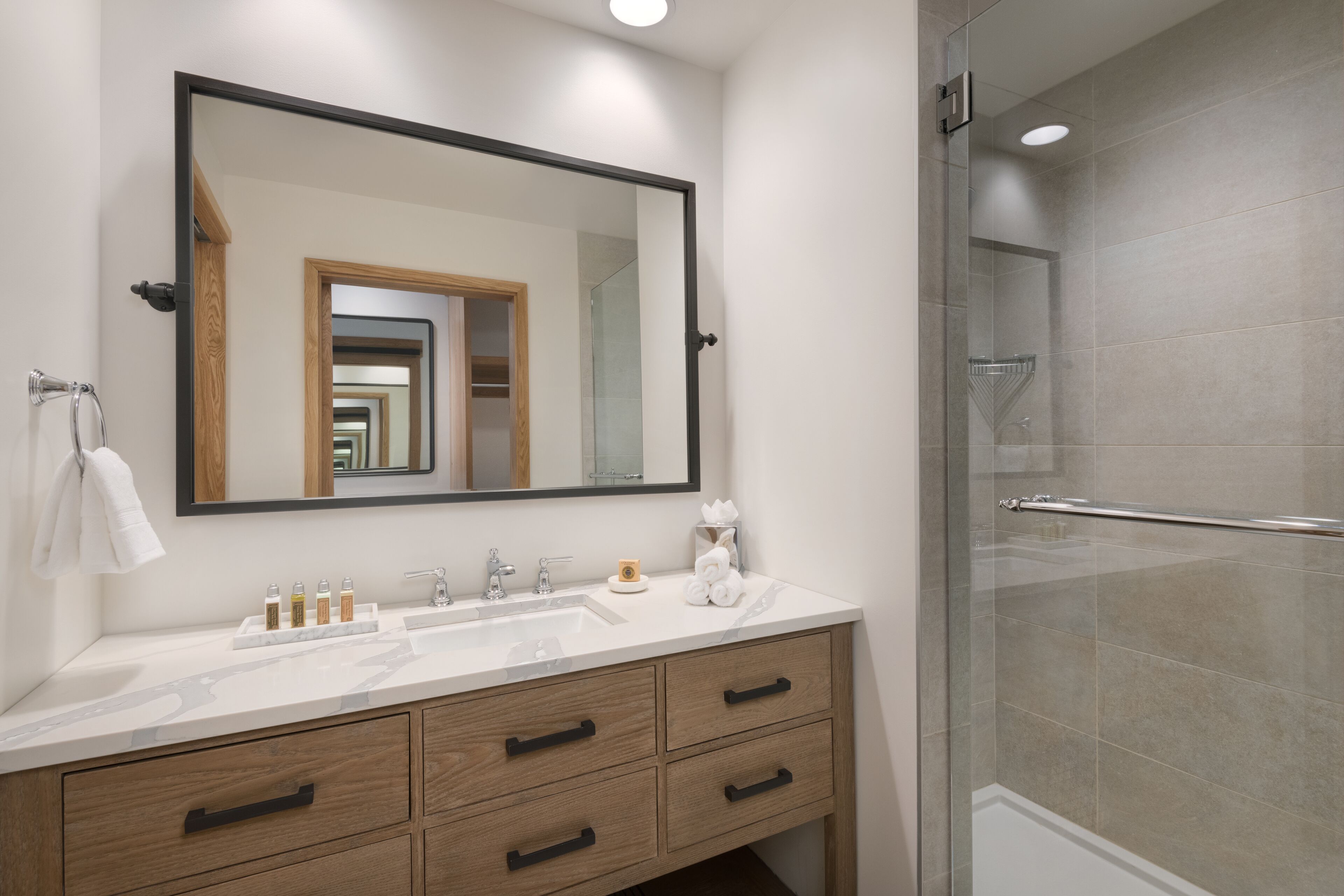 A modern bathroom with a large mirror, a sink with multiple bottles, wooden drawers, and a glass shower enclosure.
