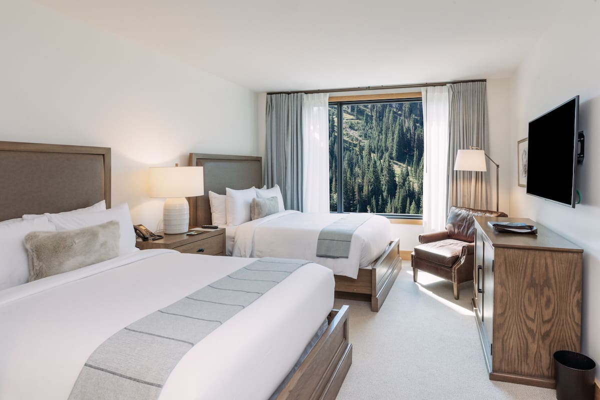 A hotel room with two beds, a desk, a chair, a TV, and a window with a forest view.