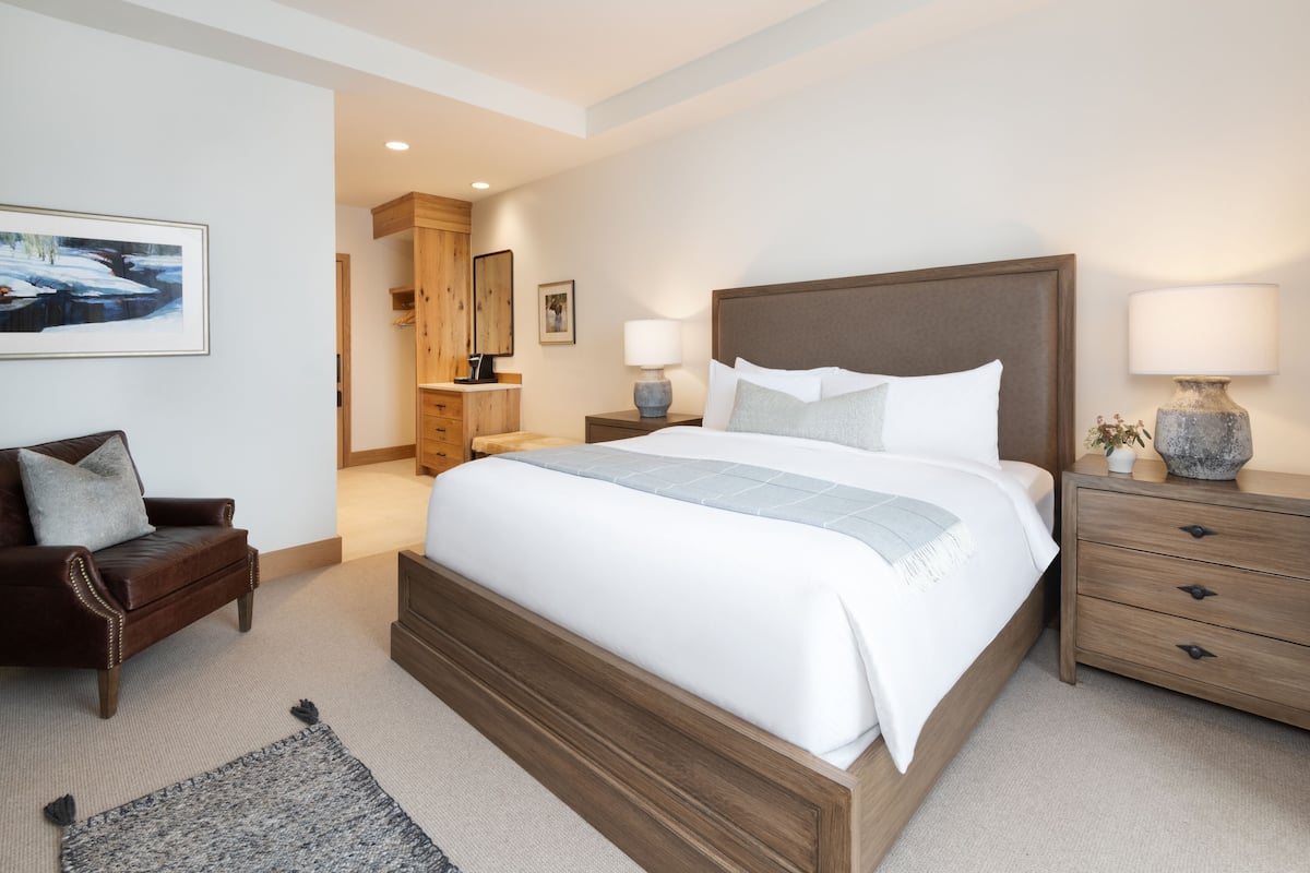 A modern hotel room with a large bed, wooden headboard, bedside tables with lamps, a leather armchair, and a framed picture on the wall.