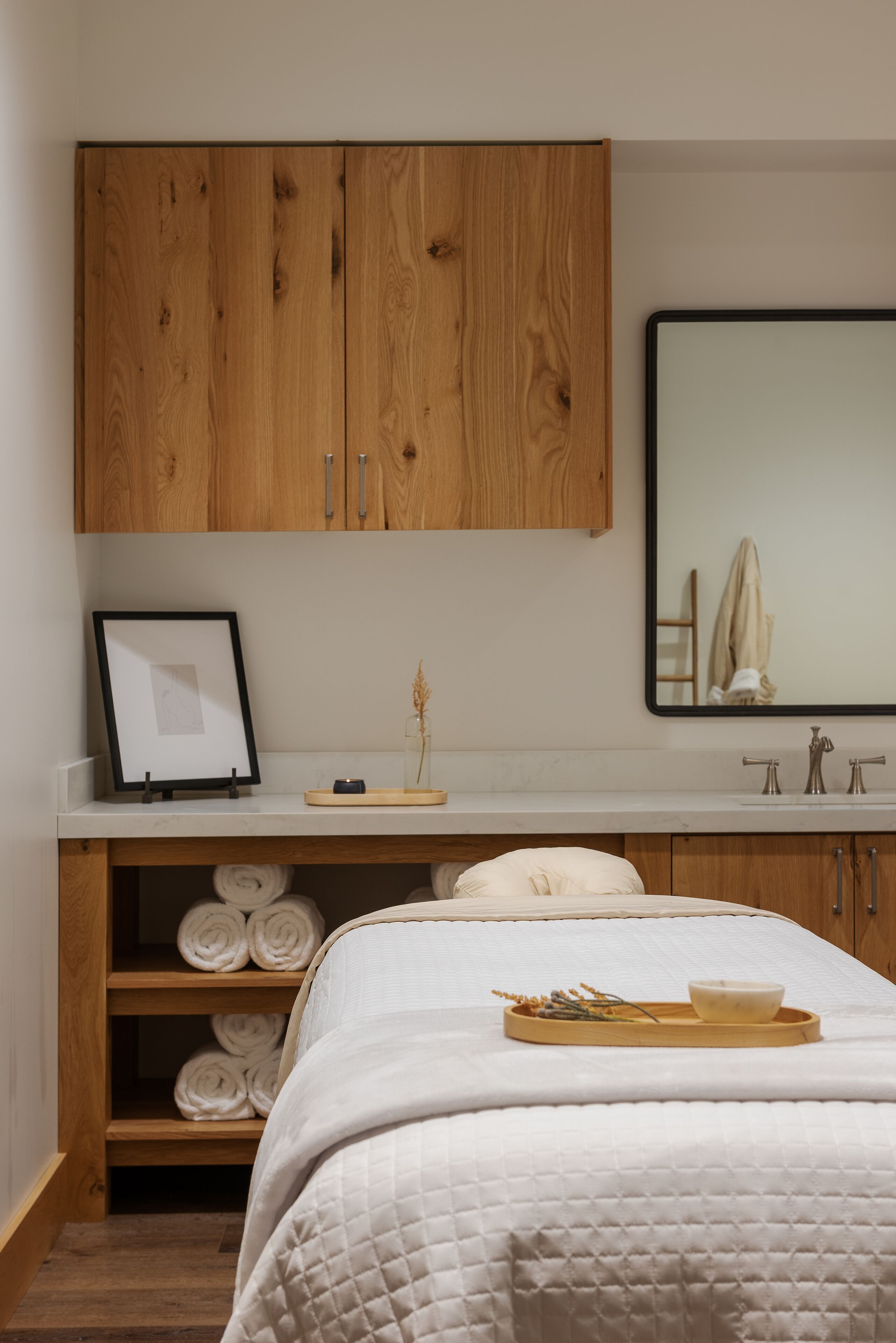 Ein Schlafzimmer mit einem Holzschrank, einem gerahmten Bild, einem Spiegel, einem Bett mit gesteppter Decke, einem Tablett mit einer Schüssel und Handtüchern auf Regalen.
