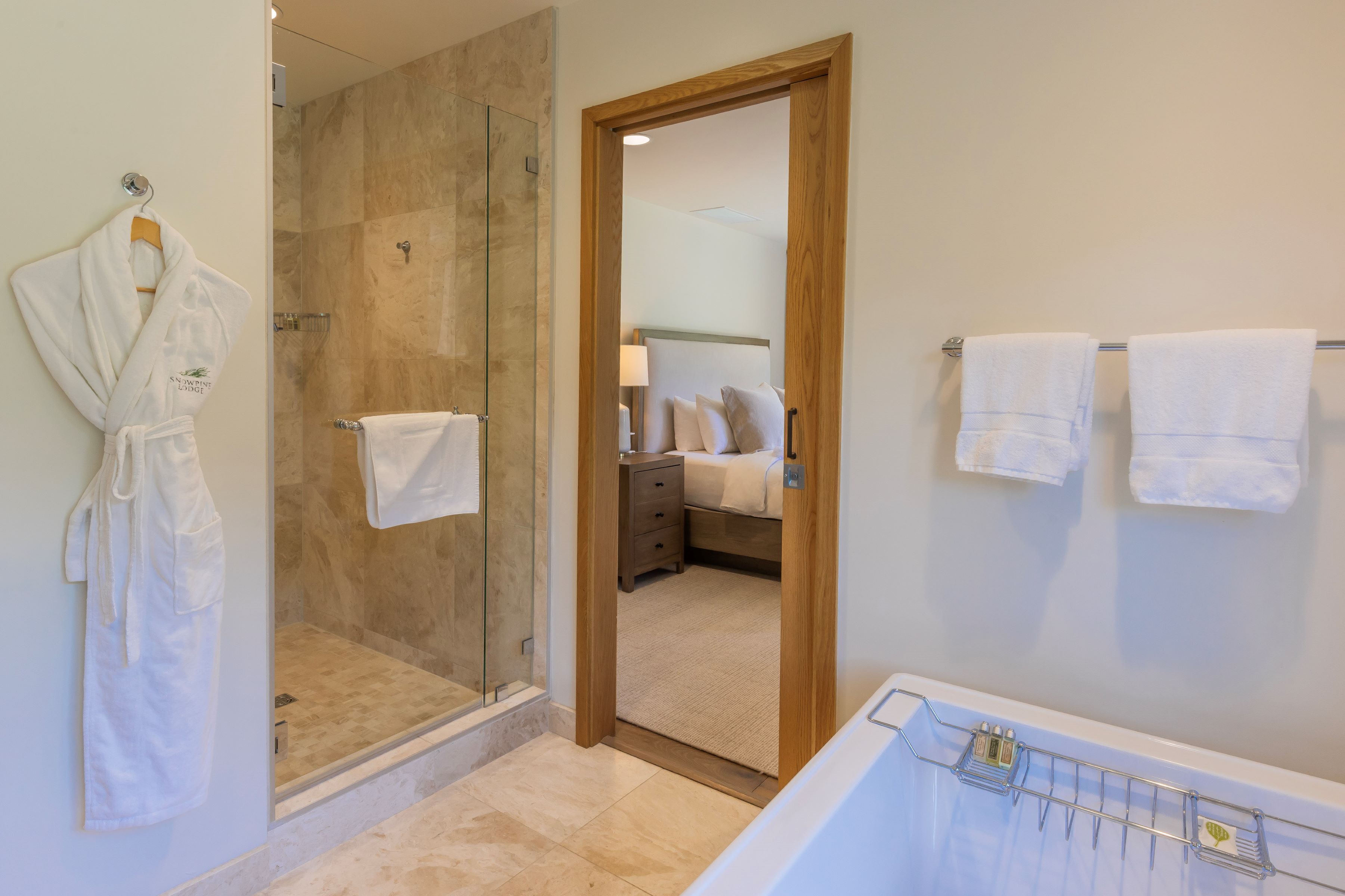 Un baño moderno con ducha de vidrio, bañera y puerta de madera que da a un dormitorio.