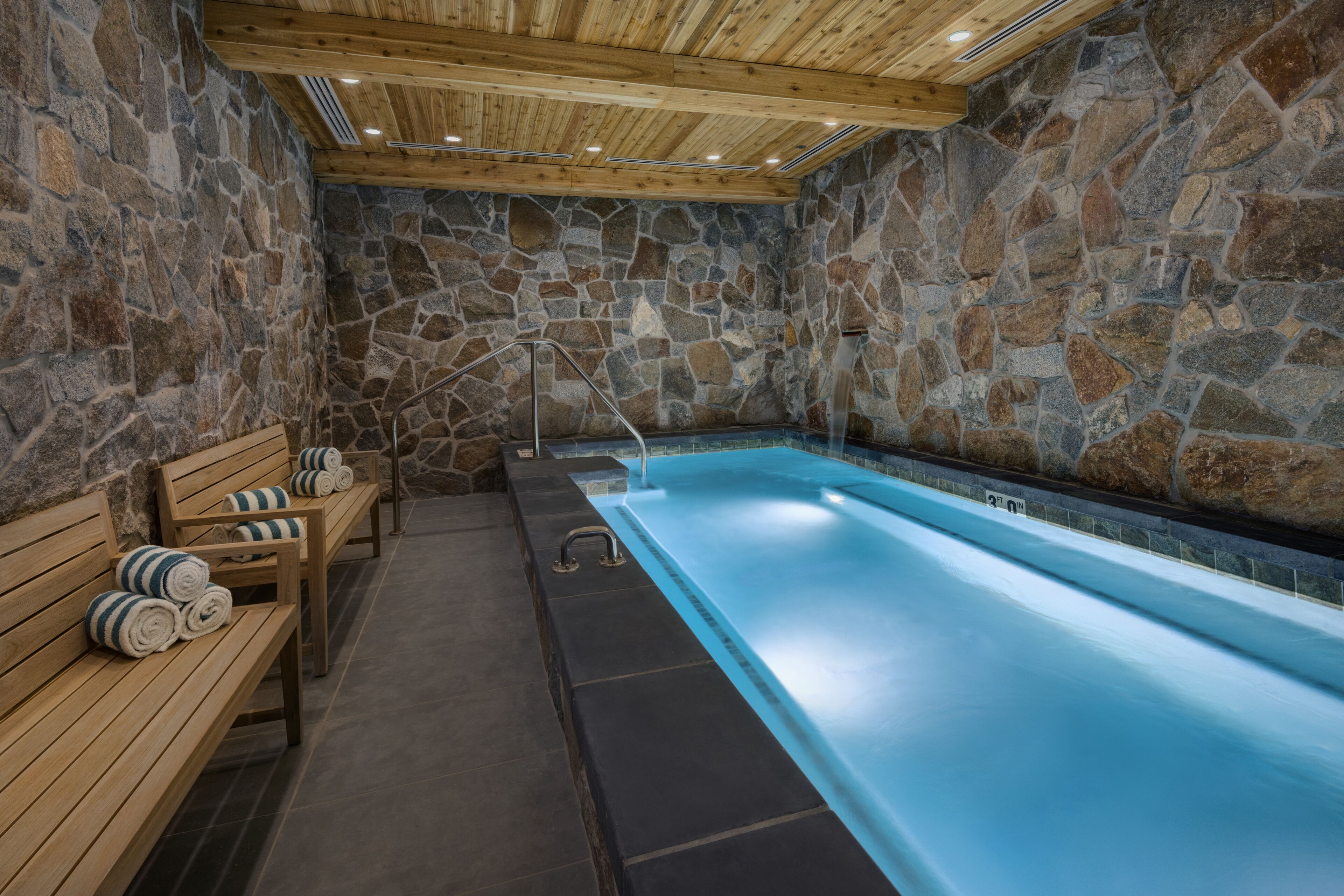 Un espace de détente aux murs de pierre, doté d’une longue et étroite piscine, de bancs en bois et d’un banc sur lequel sont pliés des serviettes.