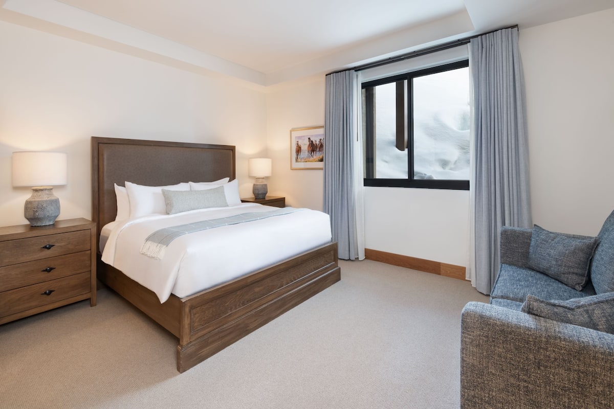 A hotel room with a large bed, a nightstand, a chair, and a window with curtains.