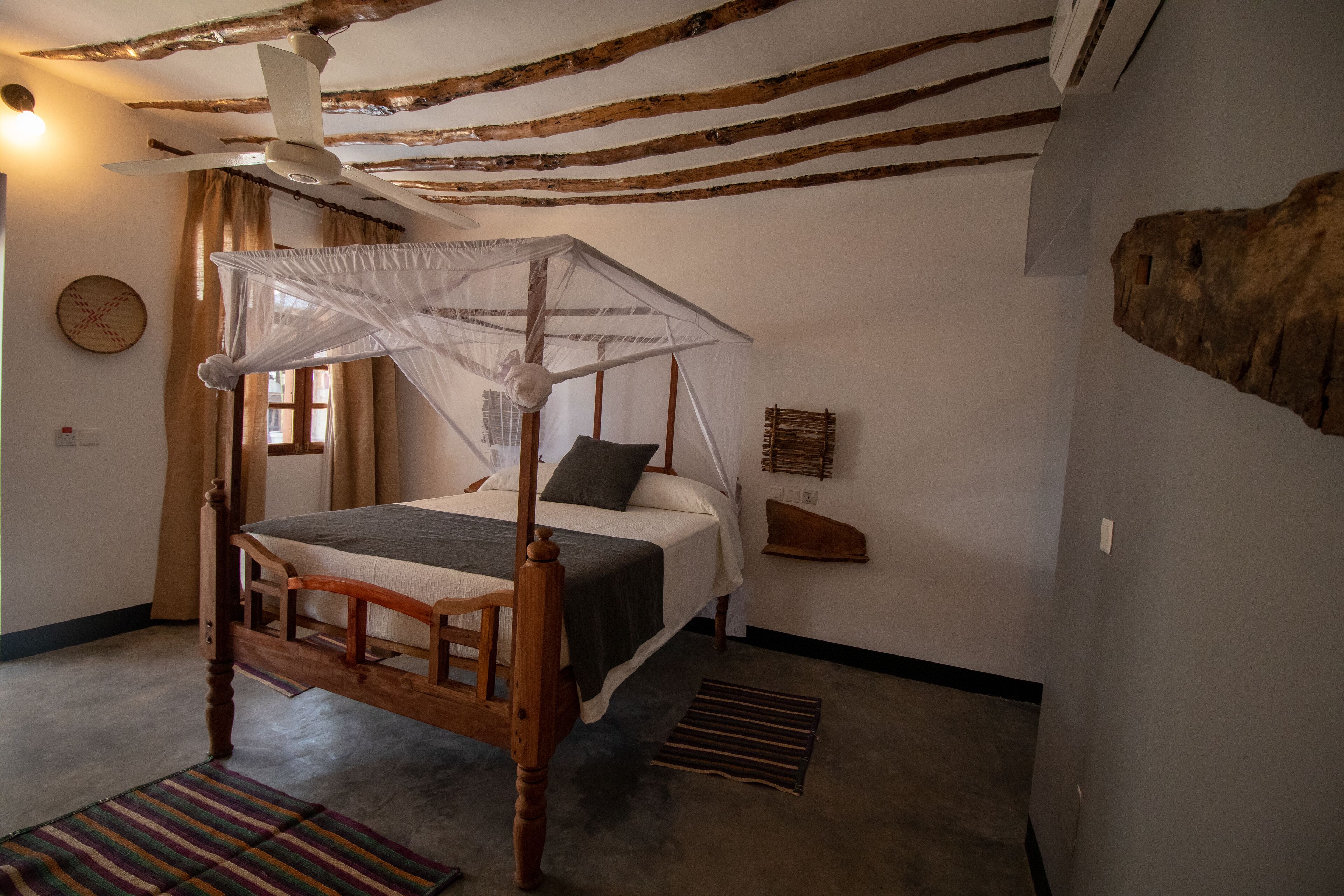Une chambre à coucher avec un lit à baldaquin, un ventilateur de plafond, une tête de lit en bois, une table de chevet, une commode, une horloge murale et une grande décoration en pierre.