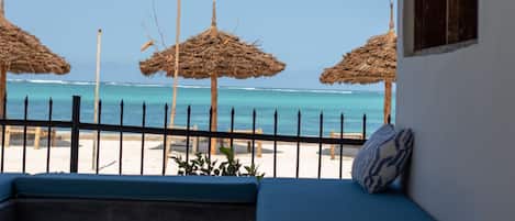 Uitzicht op een strand met rieten parasols, een hekwerk en een zitgedeelte met kussens.