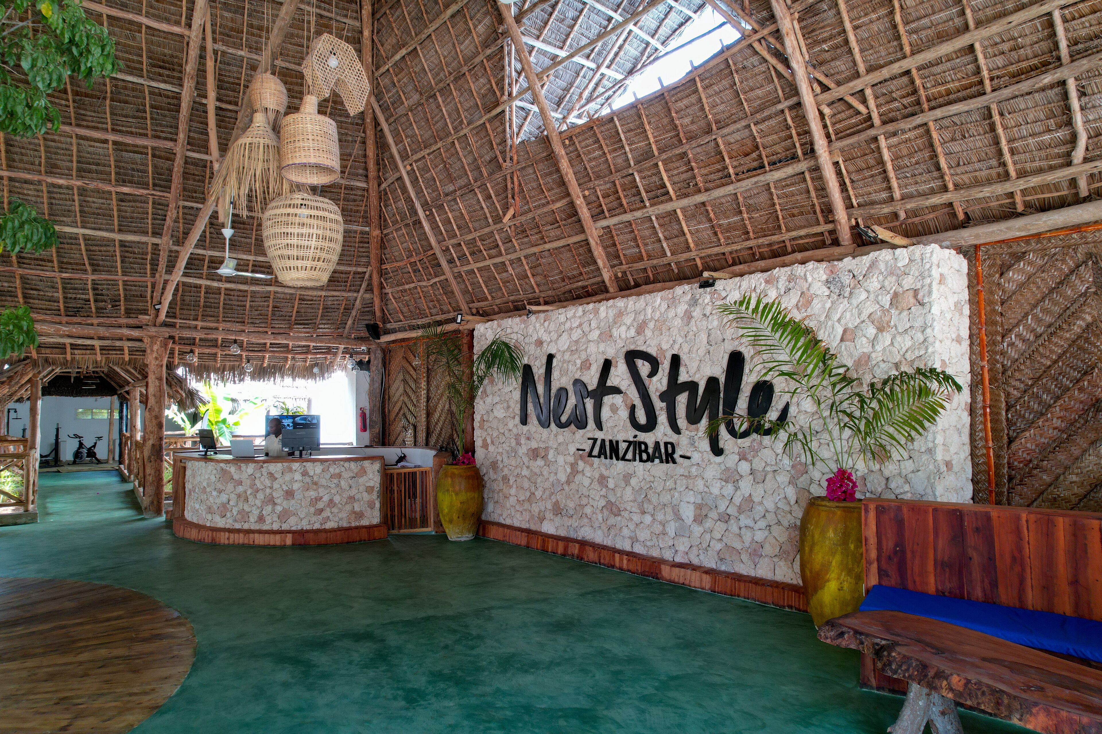 Un área de recepción con techo de paja, pared de piedra con el letrero 'Nest Style - Zanzibar', mostrador de recepción y una zona de estar con un banco.