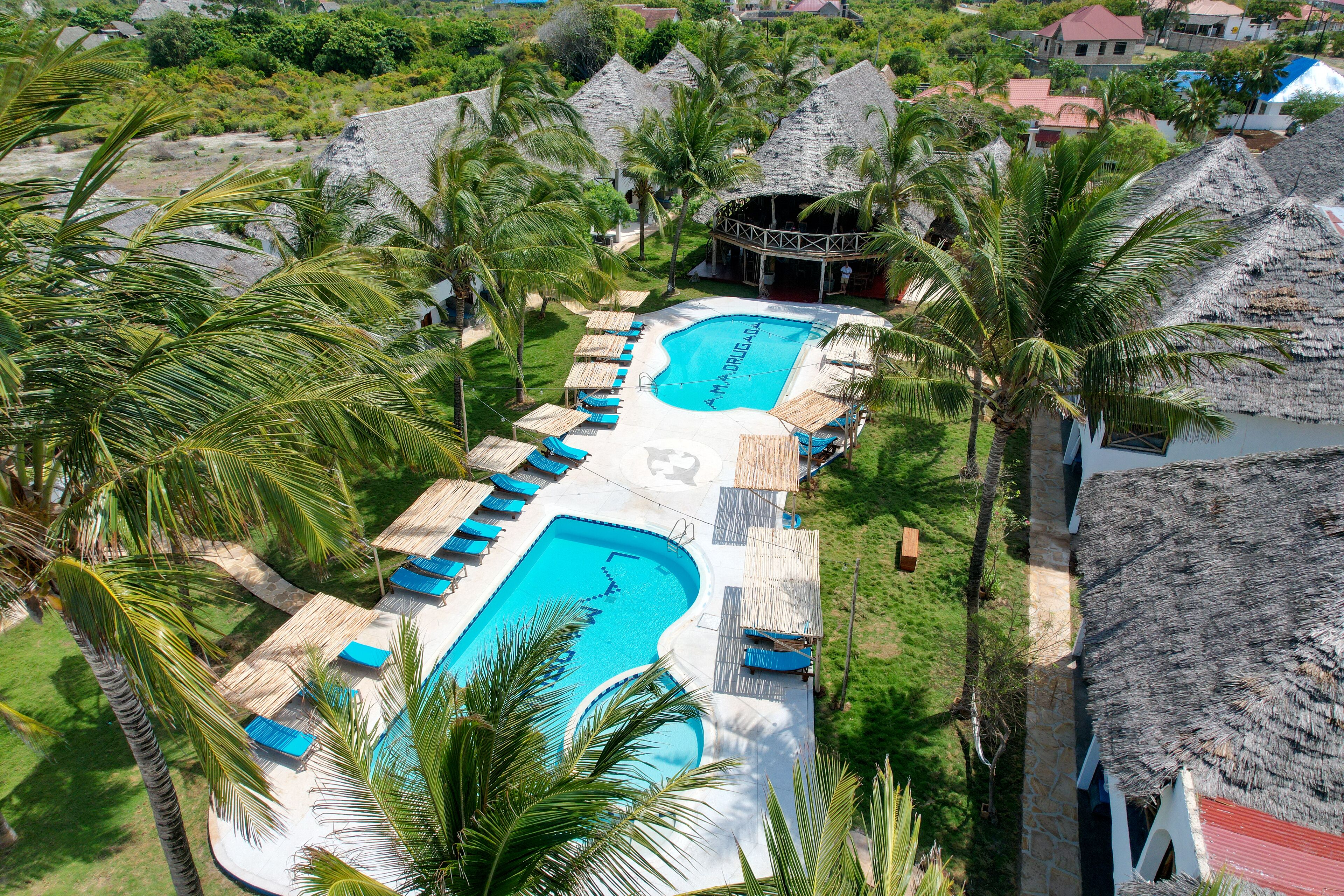 Eine Vogelperspektive zeigt ein Resort mit einem zentralen Swimmingpool, mehreren strohgedeckten Gebäuden und umliegenden Palmen.