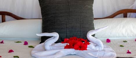 Una cama con una colcha blanca, un almohadón oscuro y una toalla doblada en forma de cisne, adornada con flores rojas.