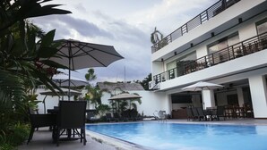 Zona de piscina con sillones de descanso y sombrillas, un edificio de varios pisos con balcones y plantas tropicales.