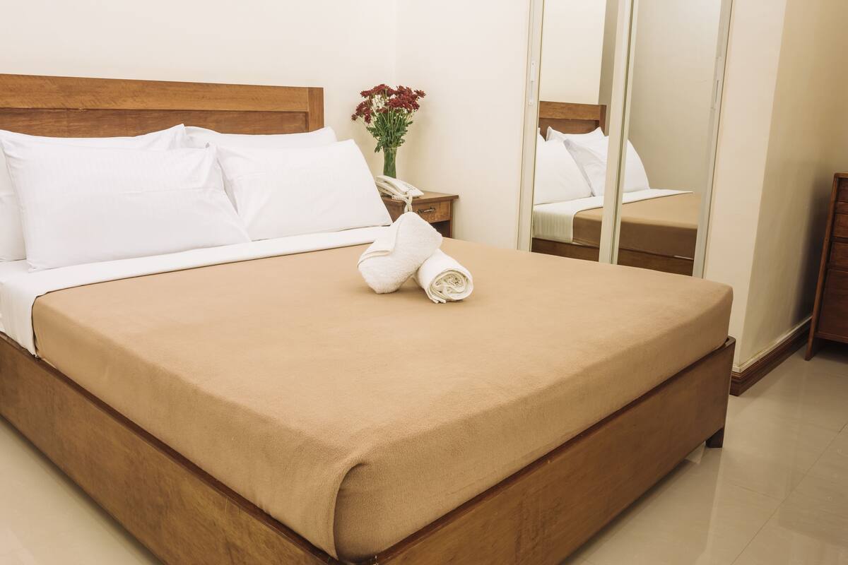 Una cama bien hecha con sábanas blancas, cabecera de madera y colcha color beige.