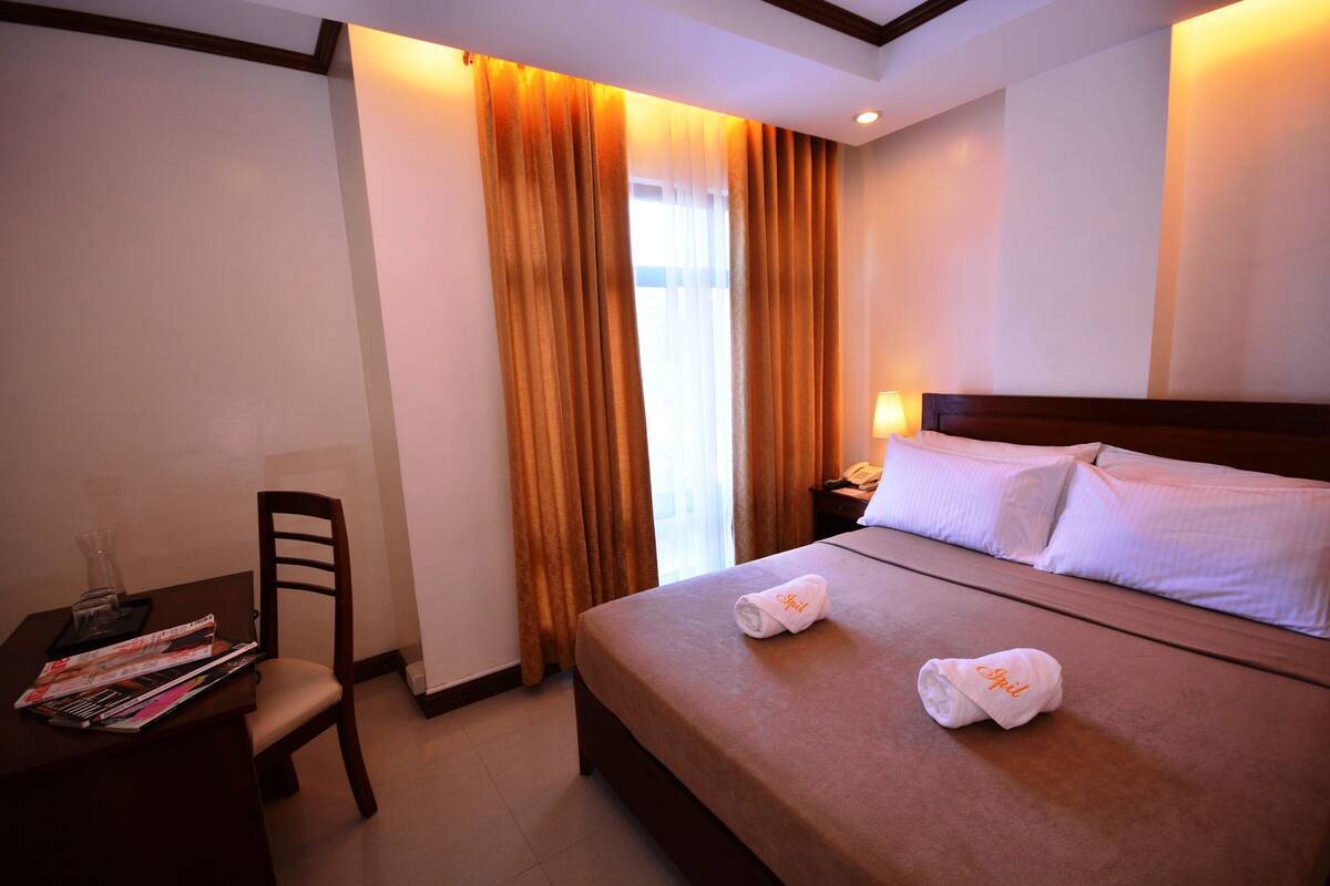 Una habitación de hotel con una cama, un escritorio con una silla, una lámpara y una ventana con cortinas.