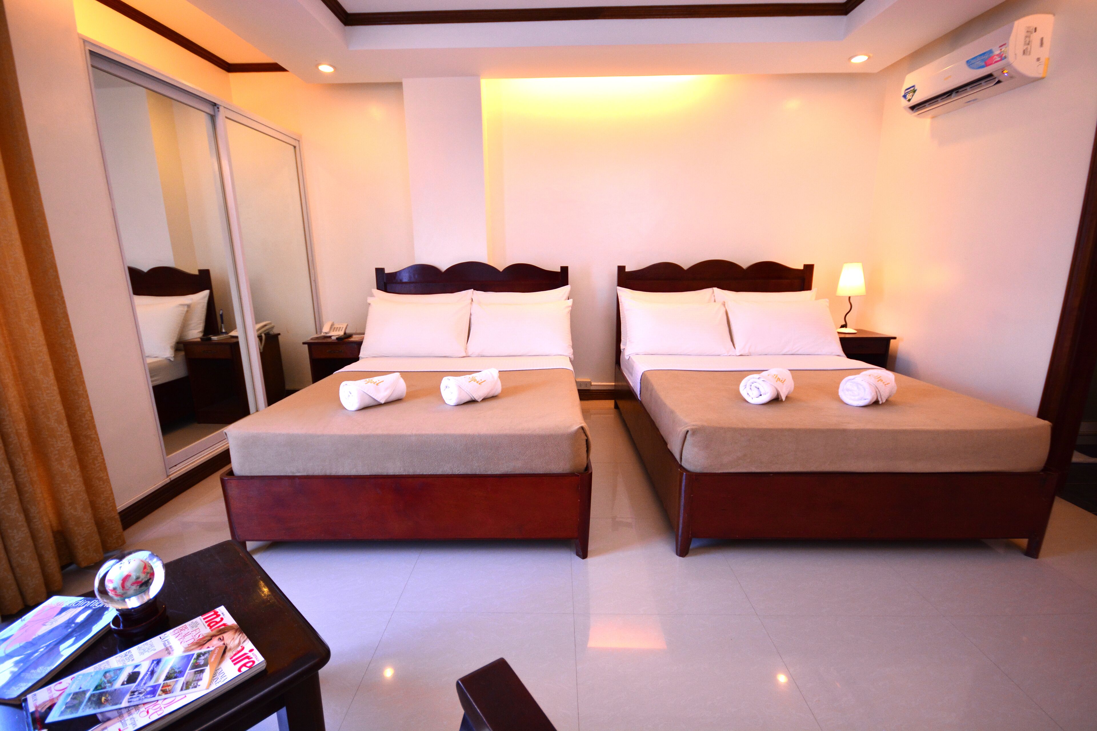 A hotel room with two beds, a nightstand with a lamp, a table with magazines, and a mirror.