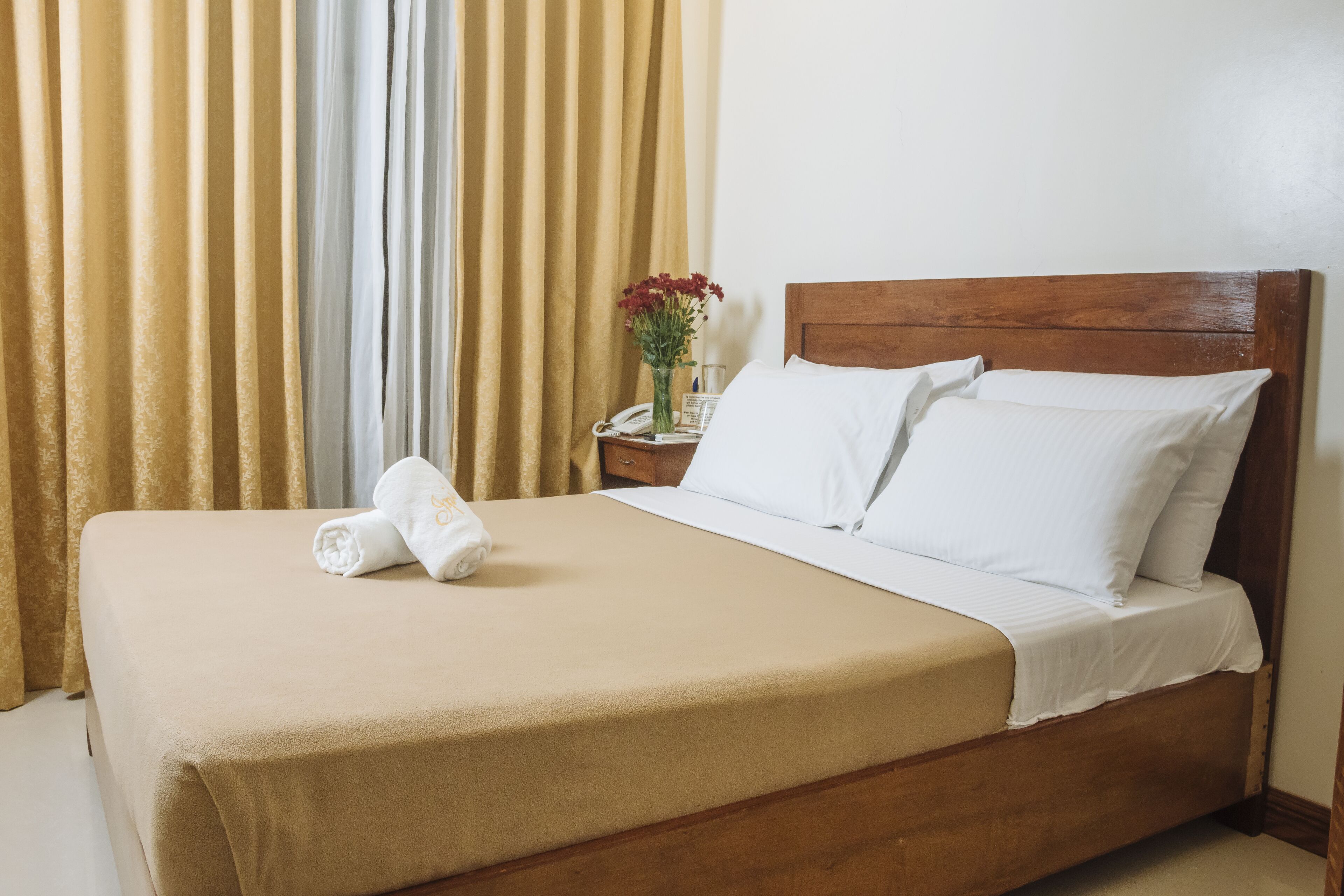A neatly made bed with white linens and a beige bedspread, a wooden headboard, and a nightstand with a vase of flowers.