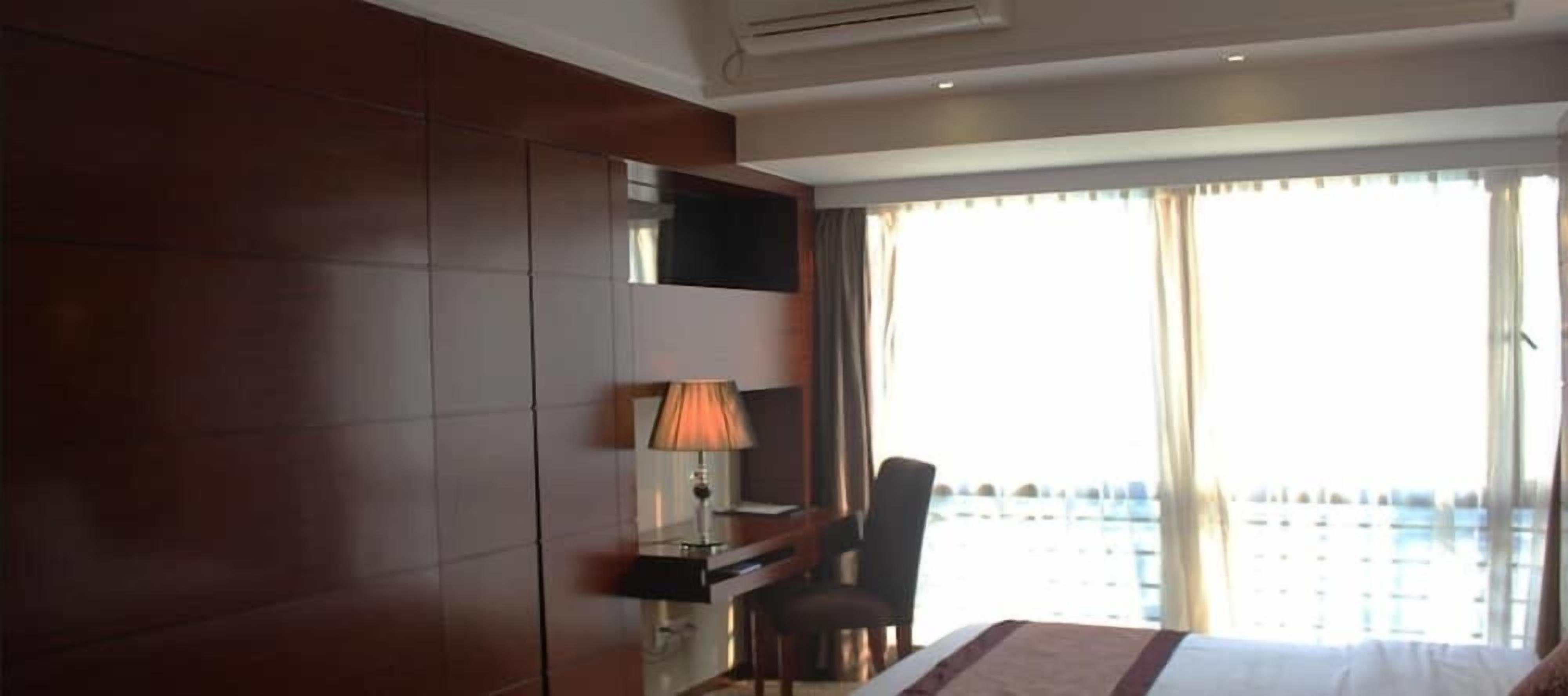 Habitación de hotel con una cama, un escritorio con lámpara, una silla y un ventanal con cortinas.