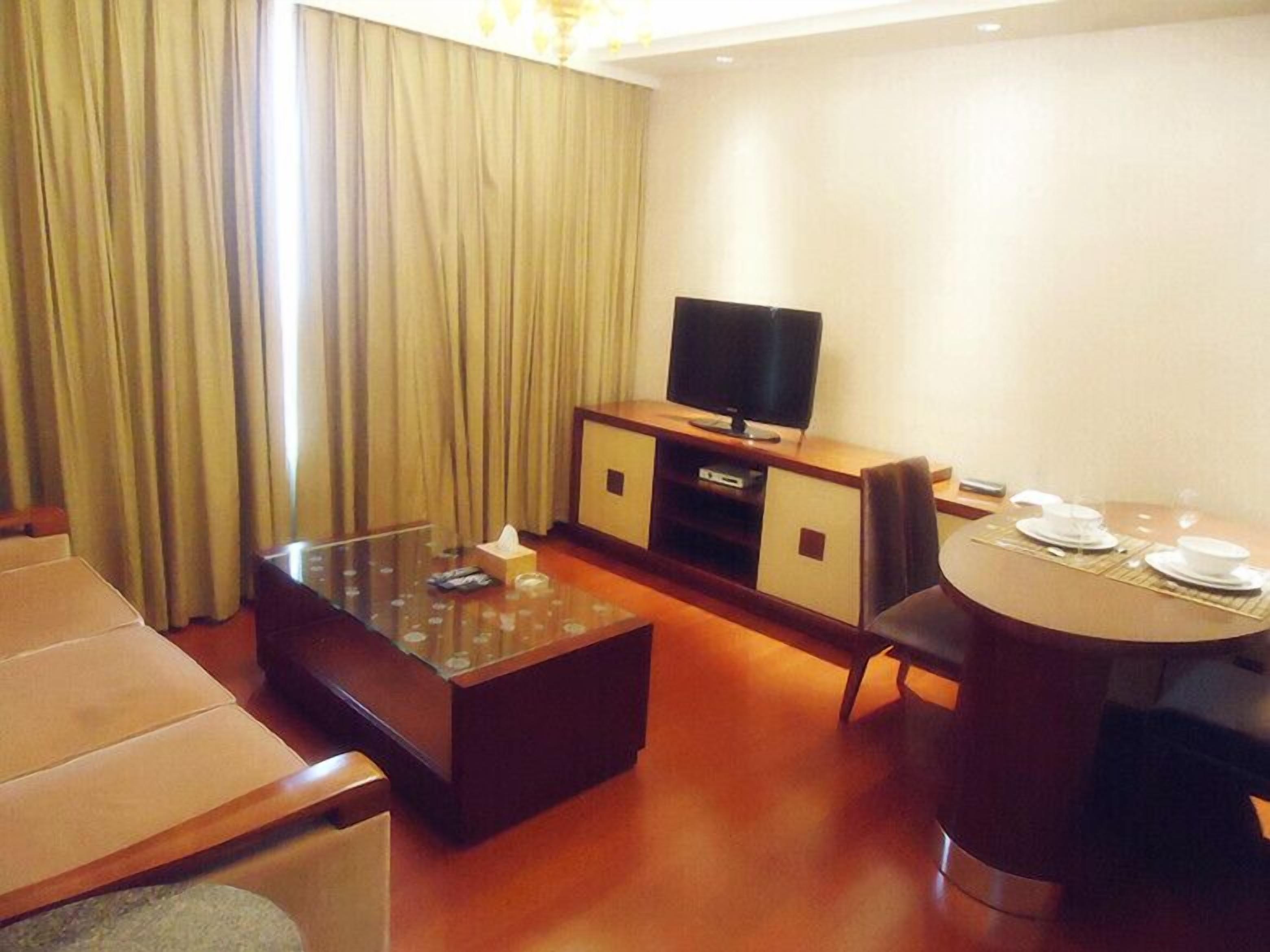 A hotel room with a sofa, a glass-top coffee table, a TV on a wooden cabinet, a dining table with plates and glasses, and a large window with curtains.