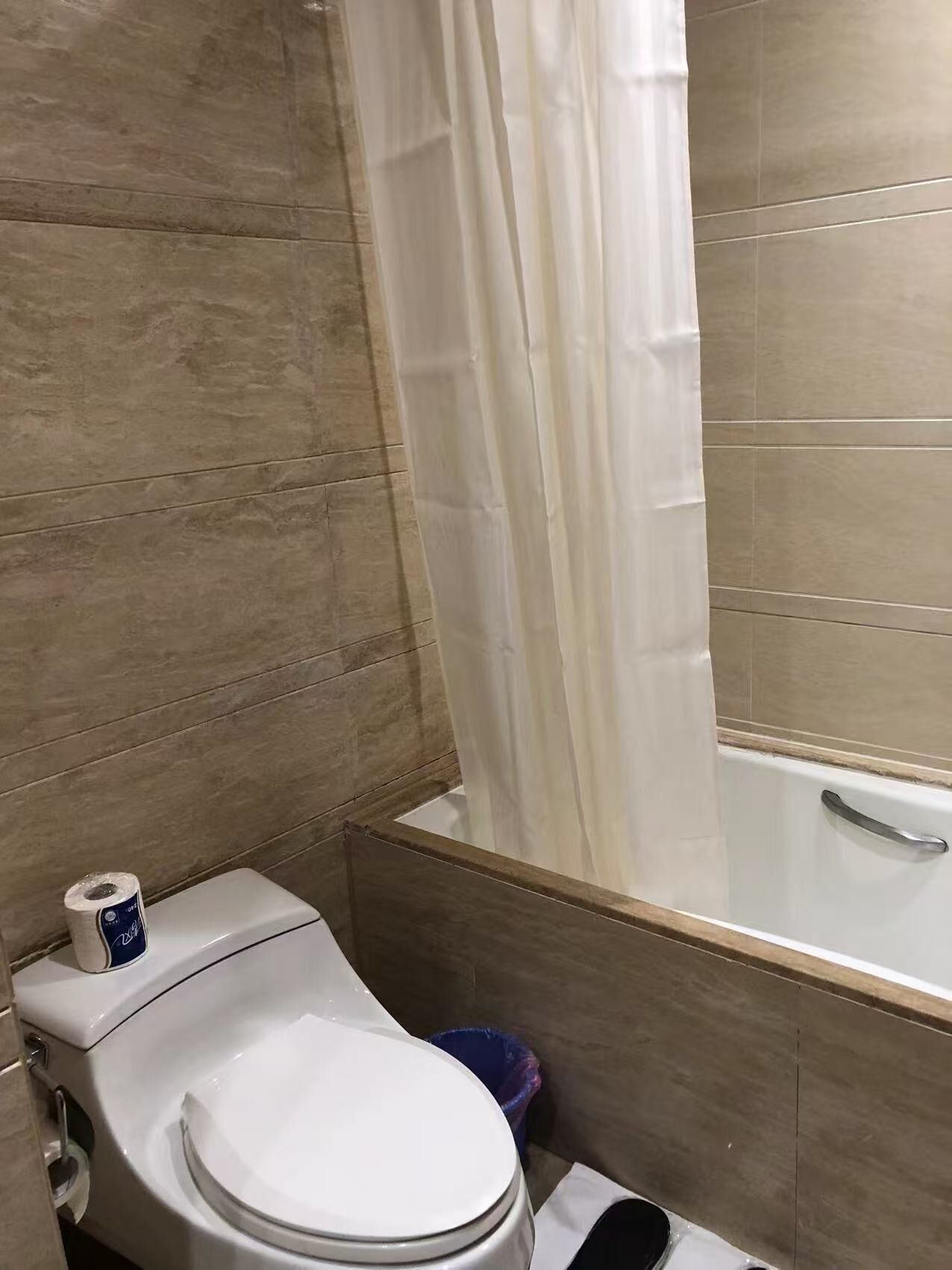 Une salle de bain avec des toilettes, un rouleau de papier toilette, une poubelle bleue et une baignoire avec un rideau de douche blanc.