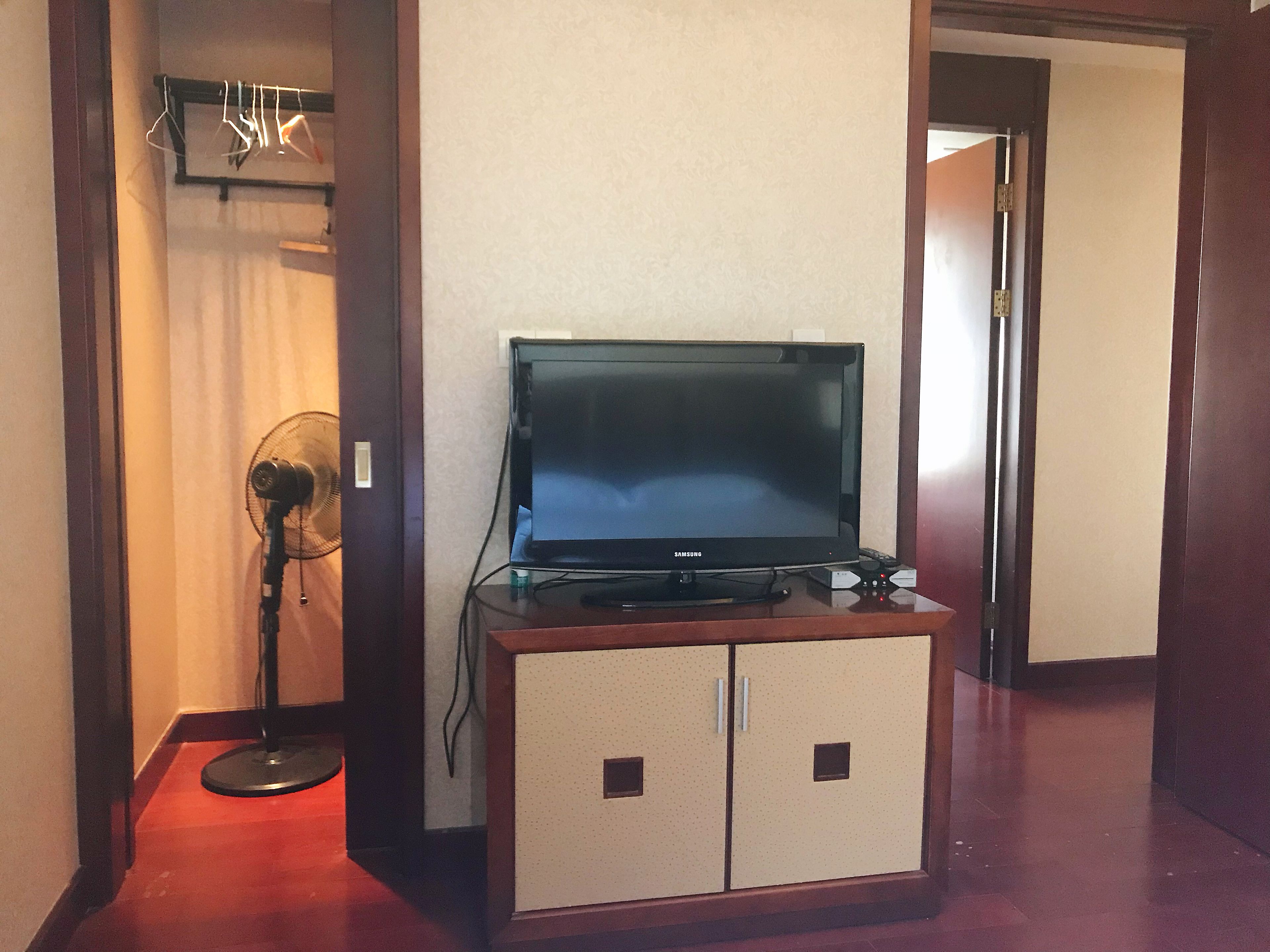 Una habitación con un televisor Samsung sobre un mueble de madera, un ventilador de pie y una puerta que da a otra habitación.
