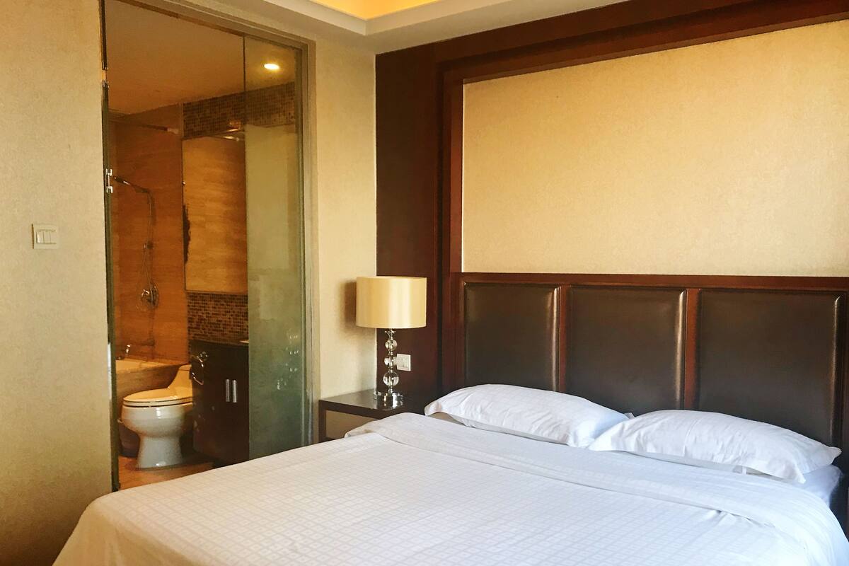 Une chambre d’hôtel avec un lit, une table de chevet avec une lampe et une salle de bain visible à travers une porte en verre.