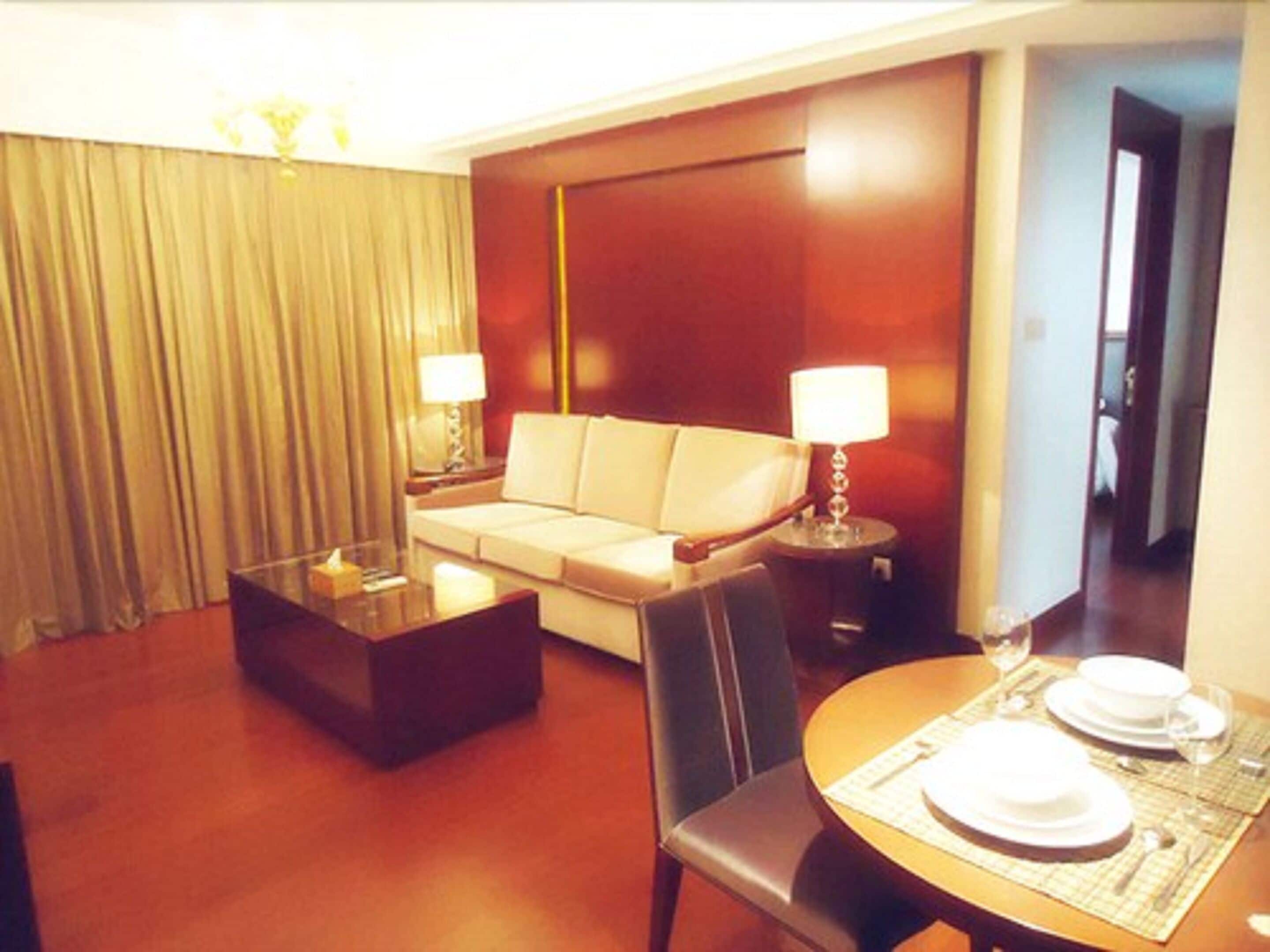 A hotel room with a sofa, a glass coffee table, a dining table set with plates and glasses, and a lamp on each side of the sofa.