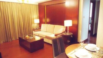 A hotel room with a sofa, a glass coffee table, a dining table set with plates and glasses, and a lamp on each side of the sofa.
