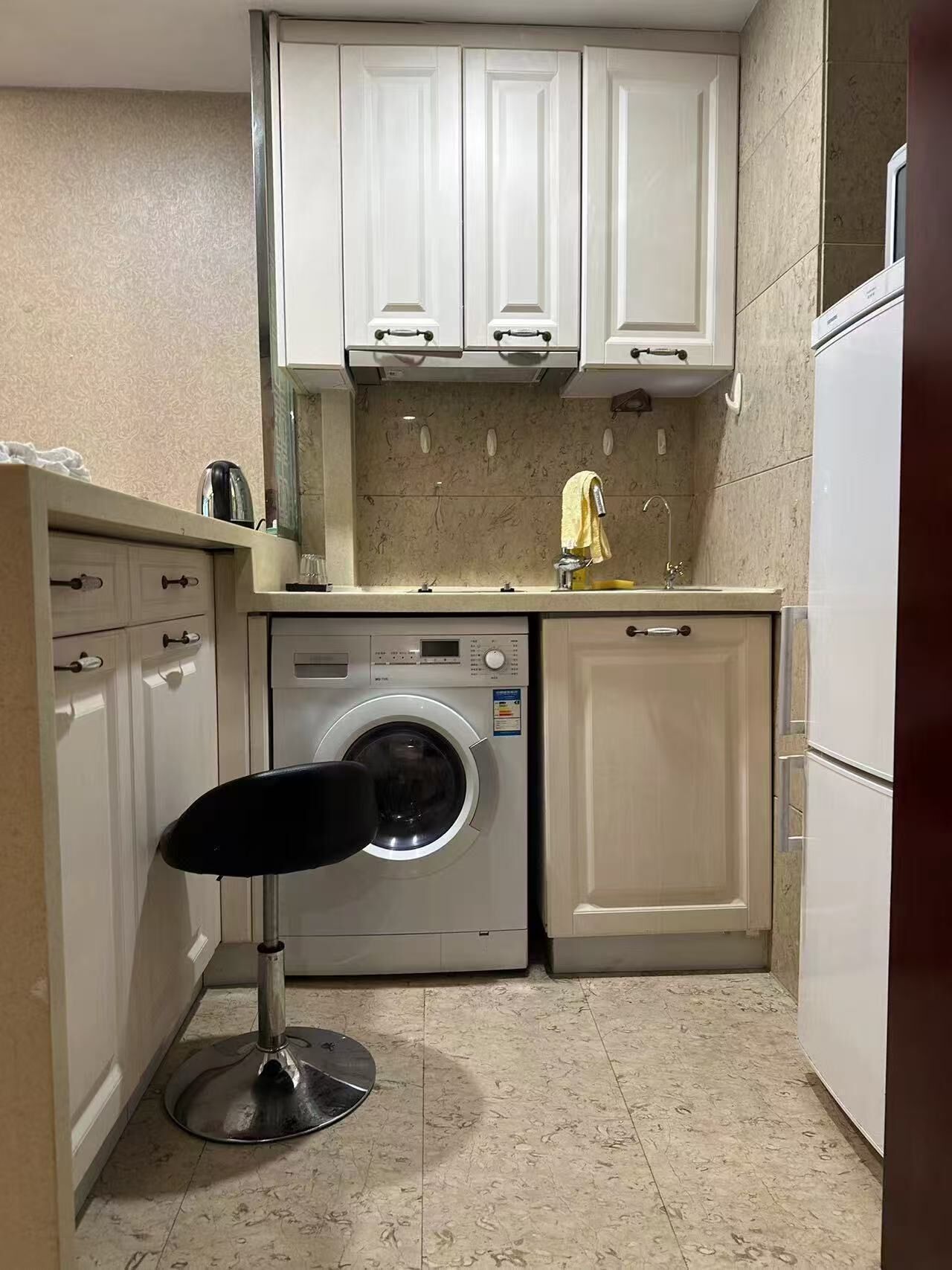 A compact kitchen with a washing machine, a sink, and a small stool.