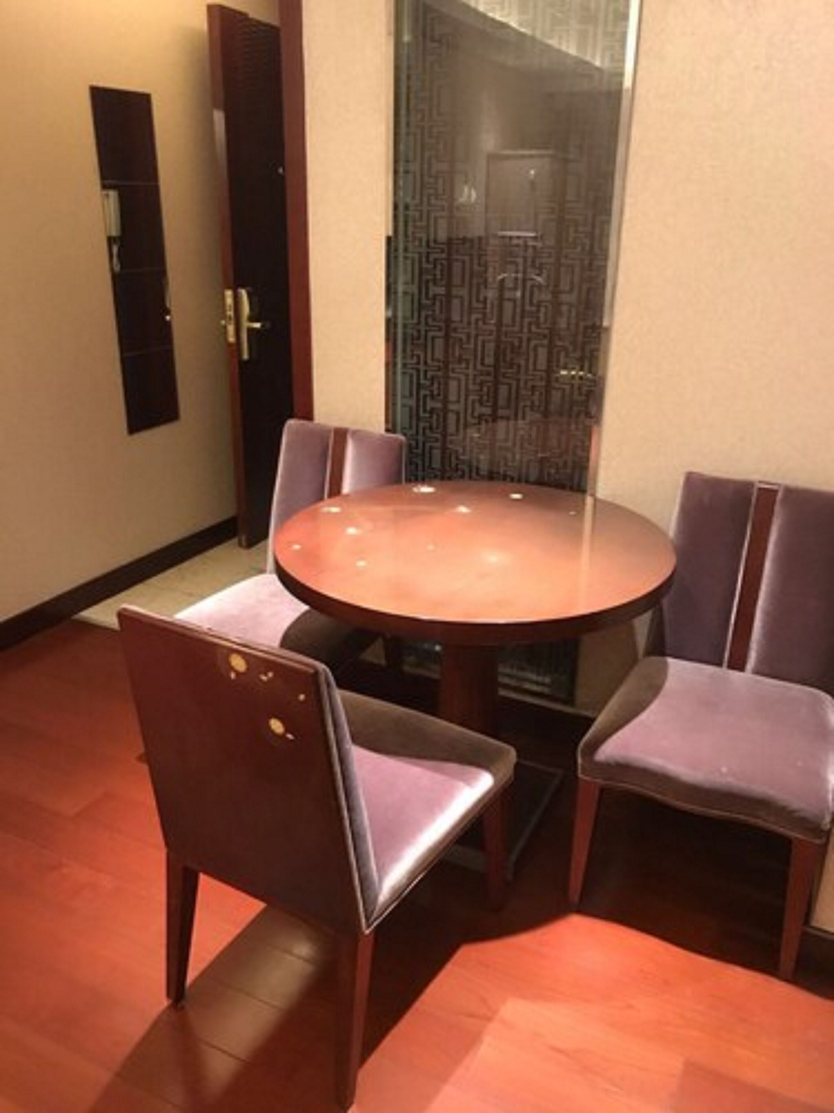 A round dining table with four chairs, a wooden floor, a door with a patterned glass panel, and a wall-mounted light switch.