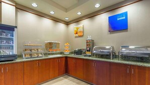 Un área de desayuno de hotel con una estación de autoservicio, un refrigerador con bebidas y un cartel en la pared.