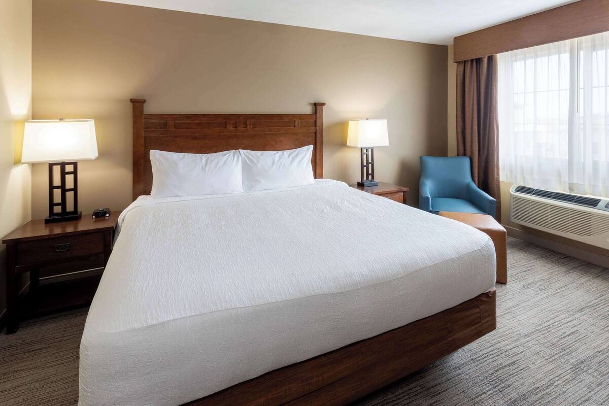 Une chambre d’hôtel avec un grand lit, deux tables de chevet avec des lampes, un fauteuil bleu et un appareil de climatisation.
