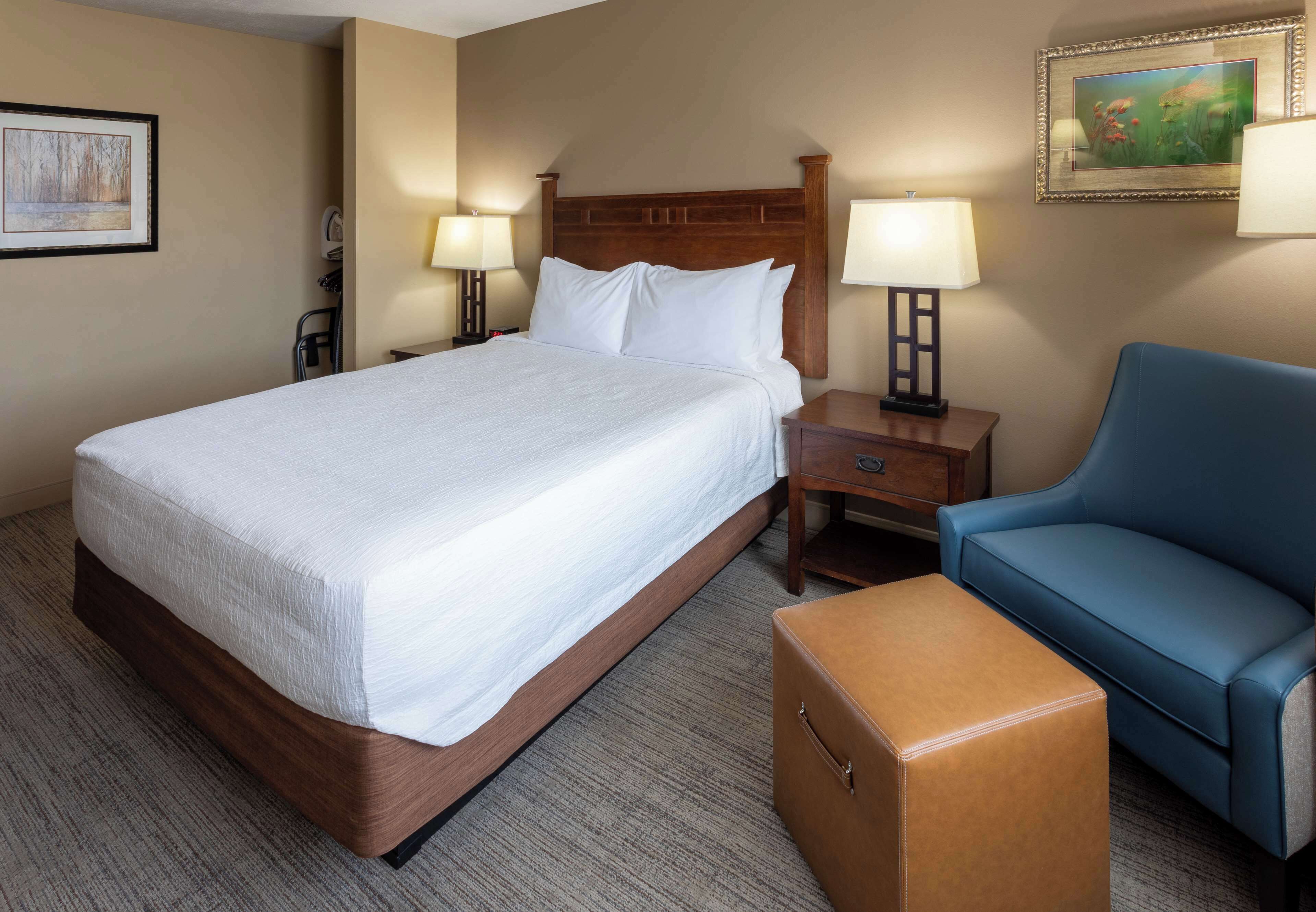 A hotel room with a large bed, bedside tables with lamps, a blue armchair, and a small wooden chest.