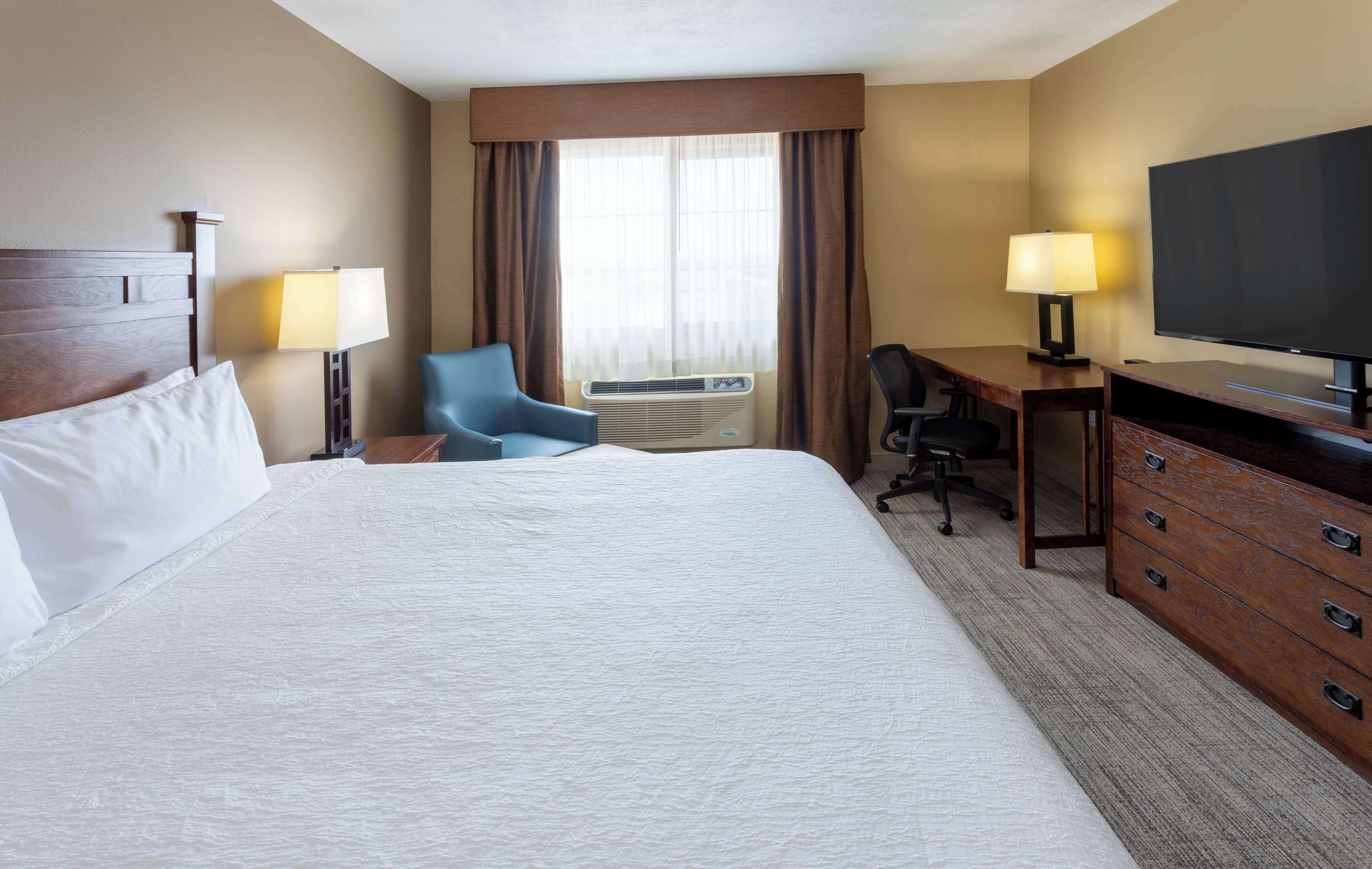 Quarto de hotel com uma cama grande, uma escrivaninha com cadeira, televisão e ar condicionado.